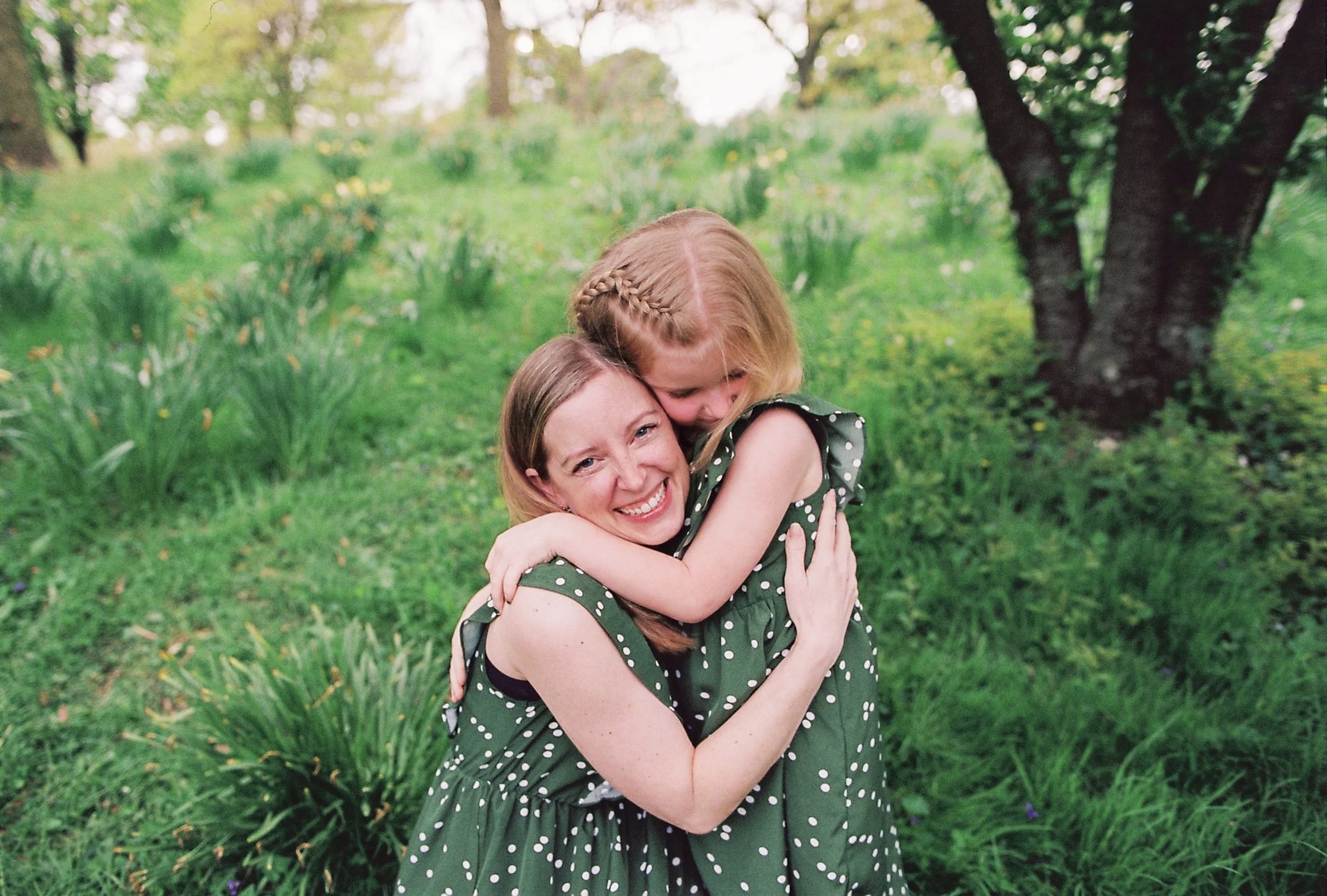 Two women hugging outdoors in a green park, smiling, with trees and bushes in the background.