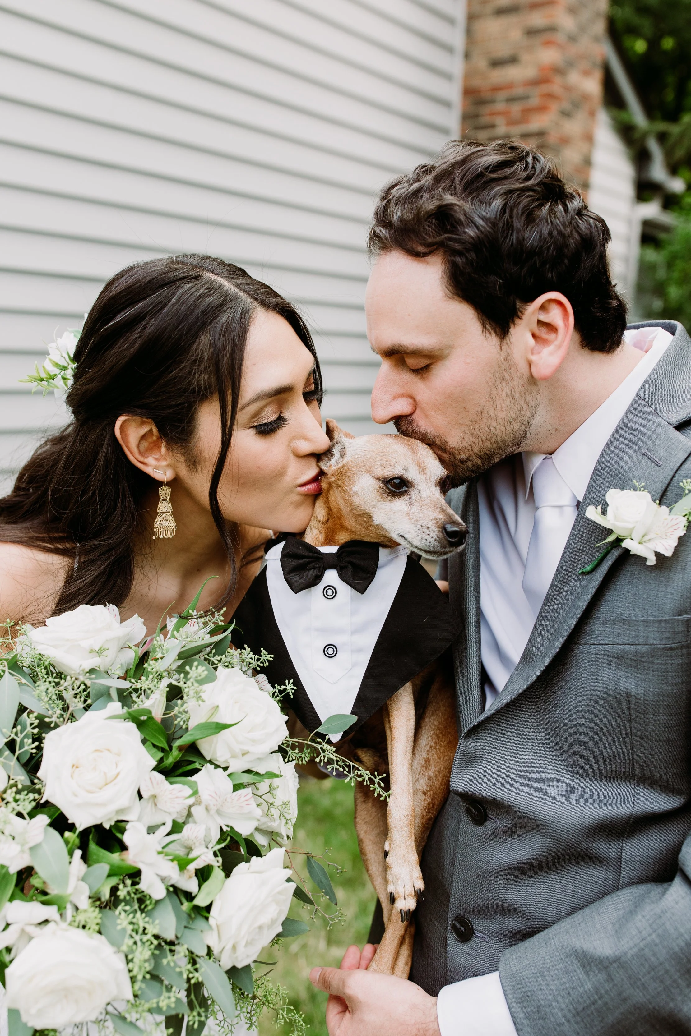 A bride and groom kissing their dog dressed in a tuxedo at a wedding.