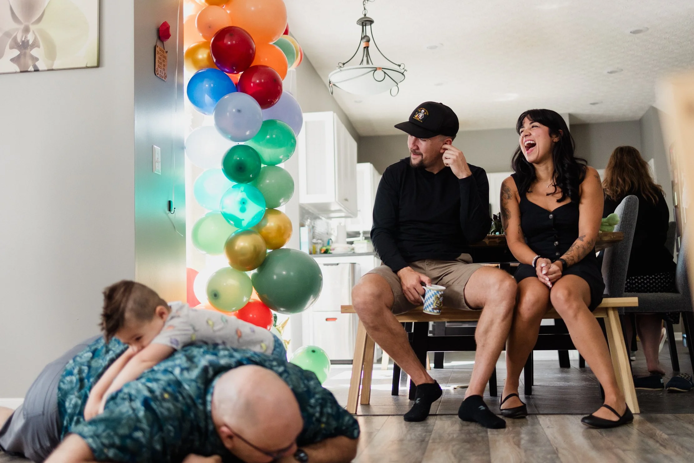 A man and woman sitting on a bench at a party, laughing. A young boy is crawling on a bald man's back on the floor. Colorful balloons are arranged in a column. People are in the background in a kitchen or dining area.