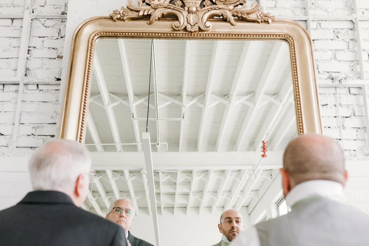 Three men in business attire are looking at themselves in a large ornate mirror with a gold frame, in a white, industrial-style room.