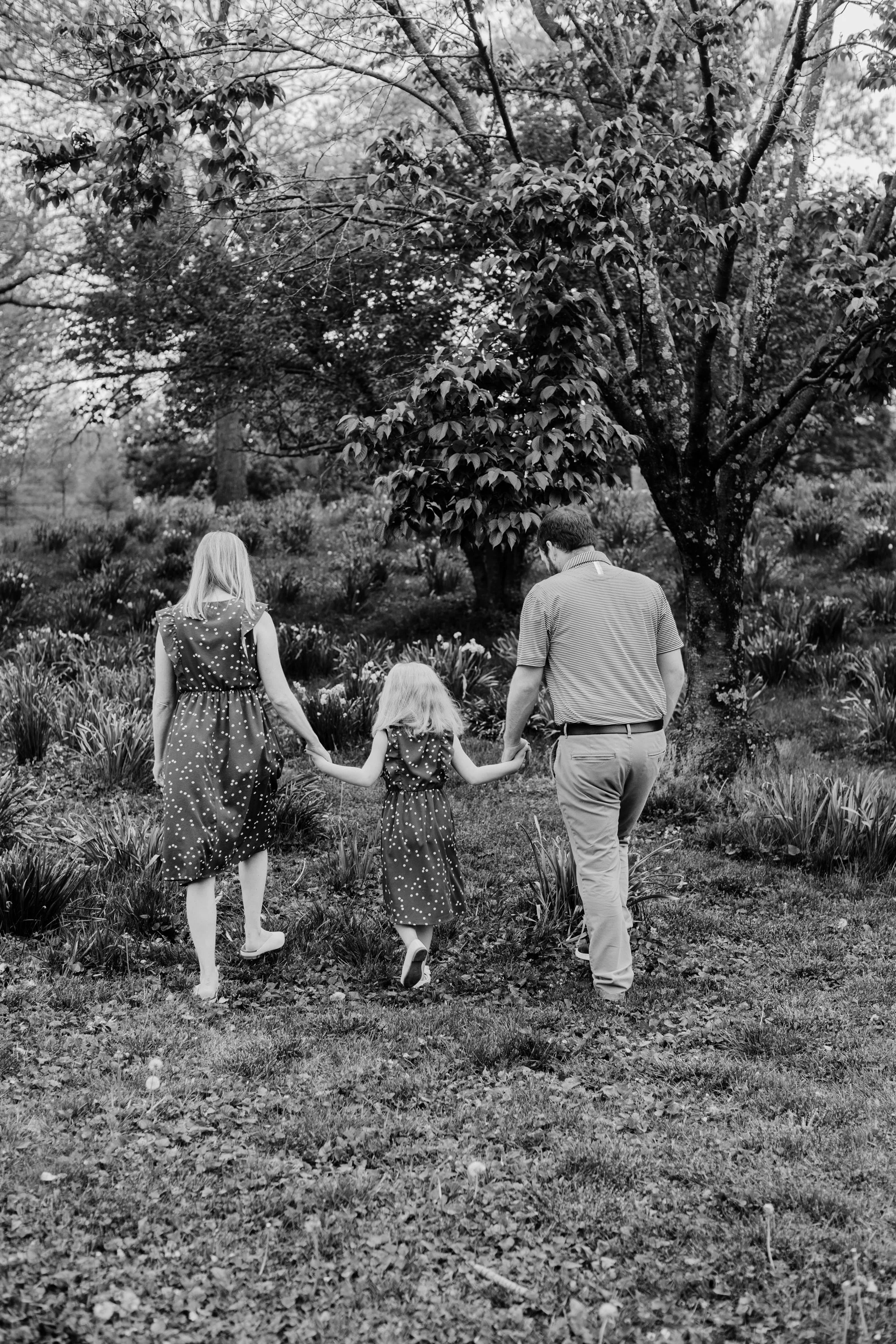 A family of three walking through a park with trees and plants, holding hands, seen from behind.