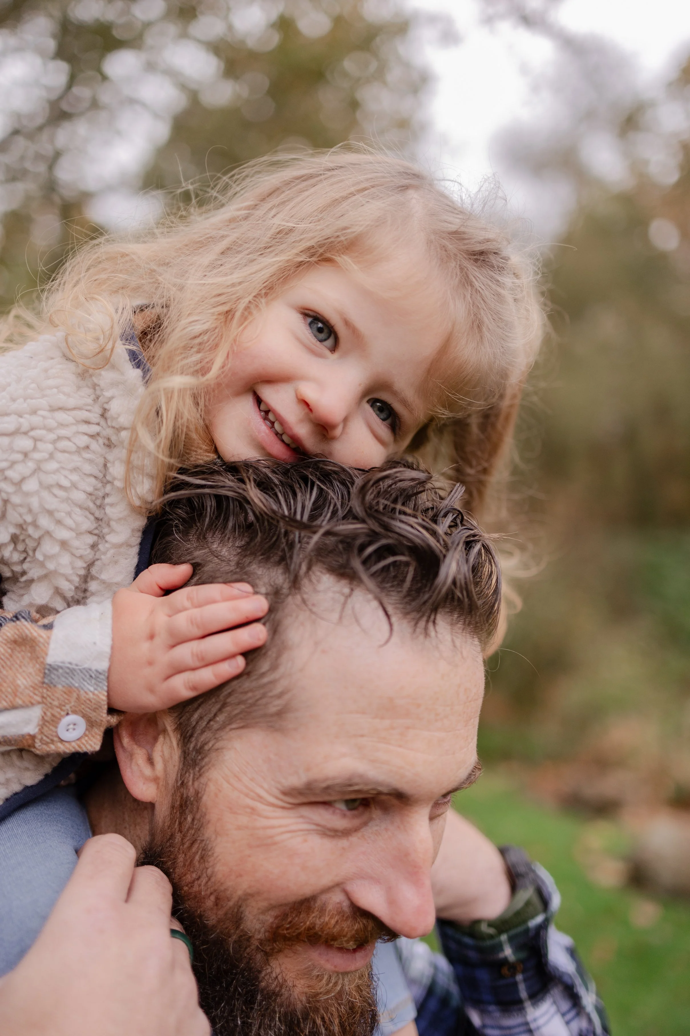 A smiling little girl with blonde hair riding on the shoulders of a man with a beard, outdoors with a blurred background of trees.