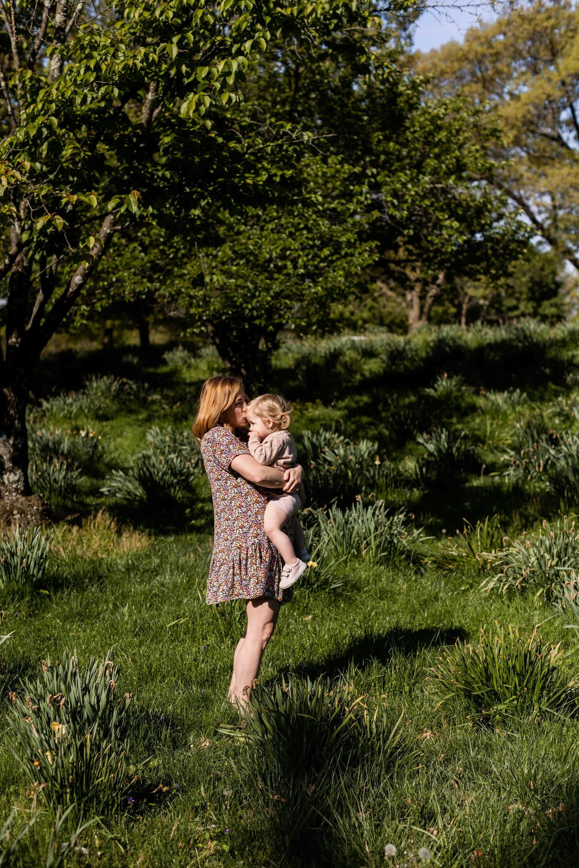 A woman holding a young girl in a green outdoor park with trees and plants in the background on a sunny day.