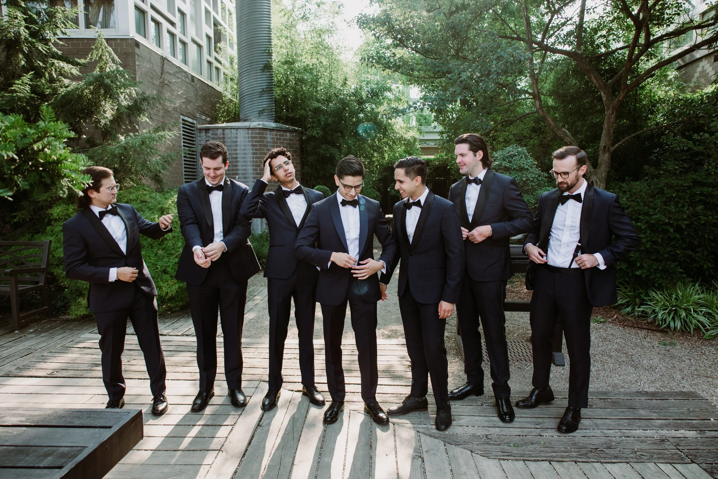 Group of seven men in tuxedos standing outdoors on a wooden deck with greenery in the background.