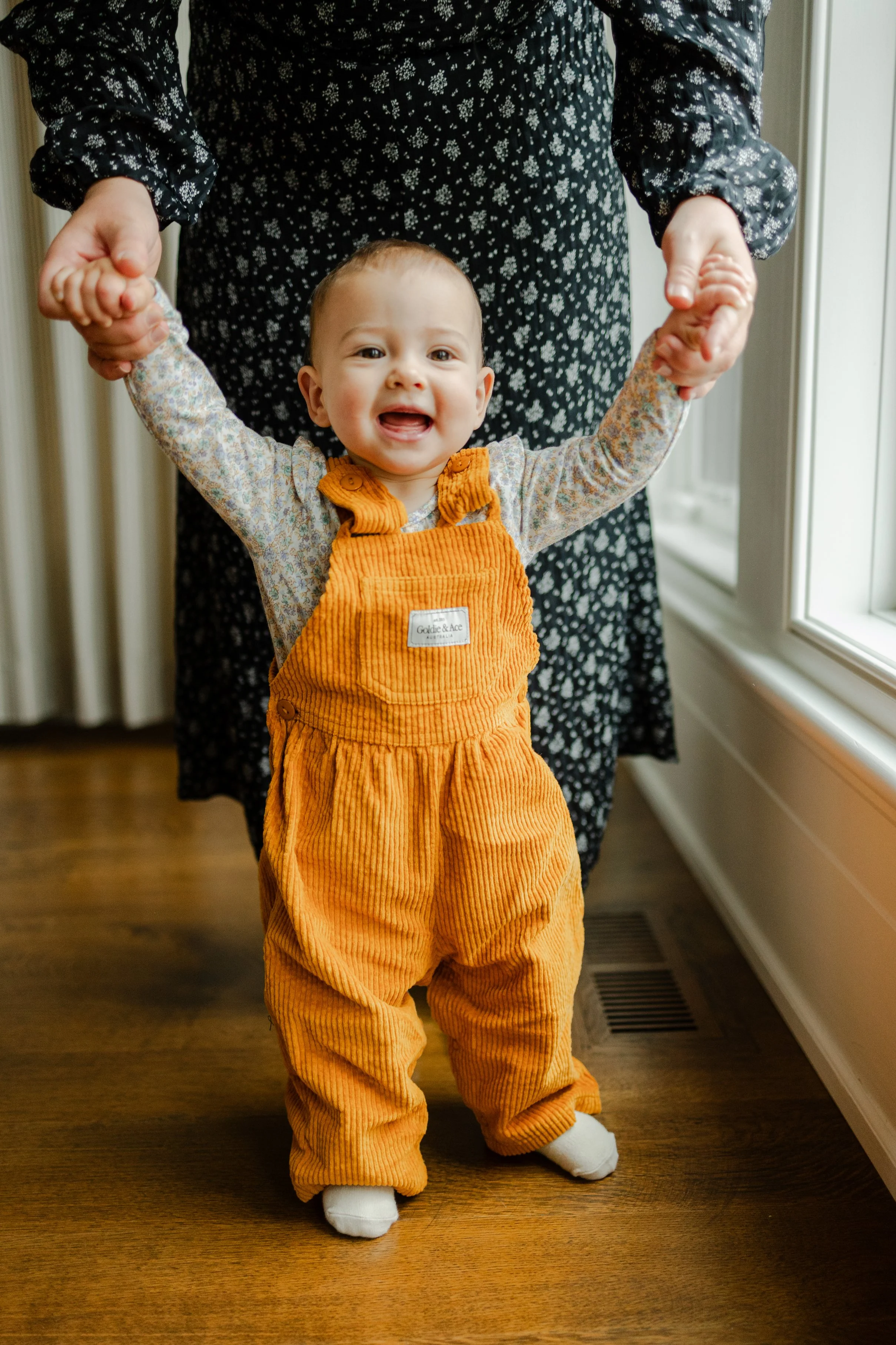 A smiling toddler in orange corduroy overalls standing on a wooden floor, holding an adult's hands for support on either side, by a window.