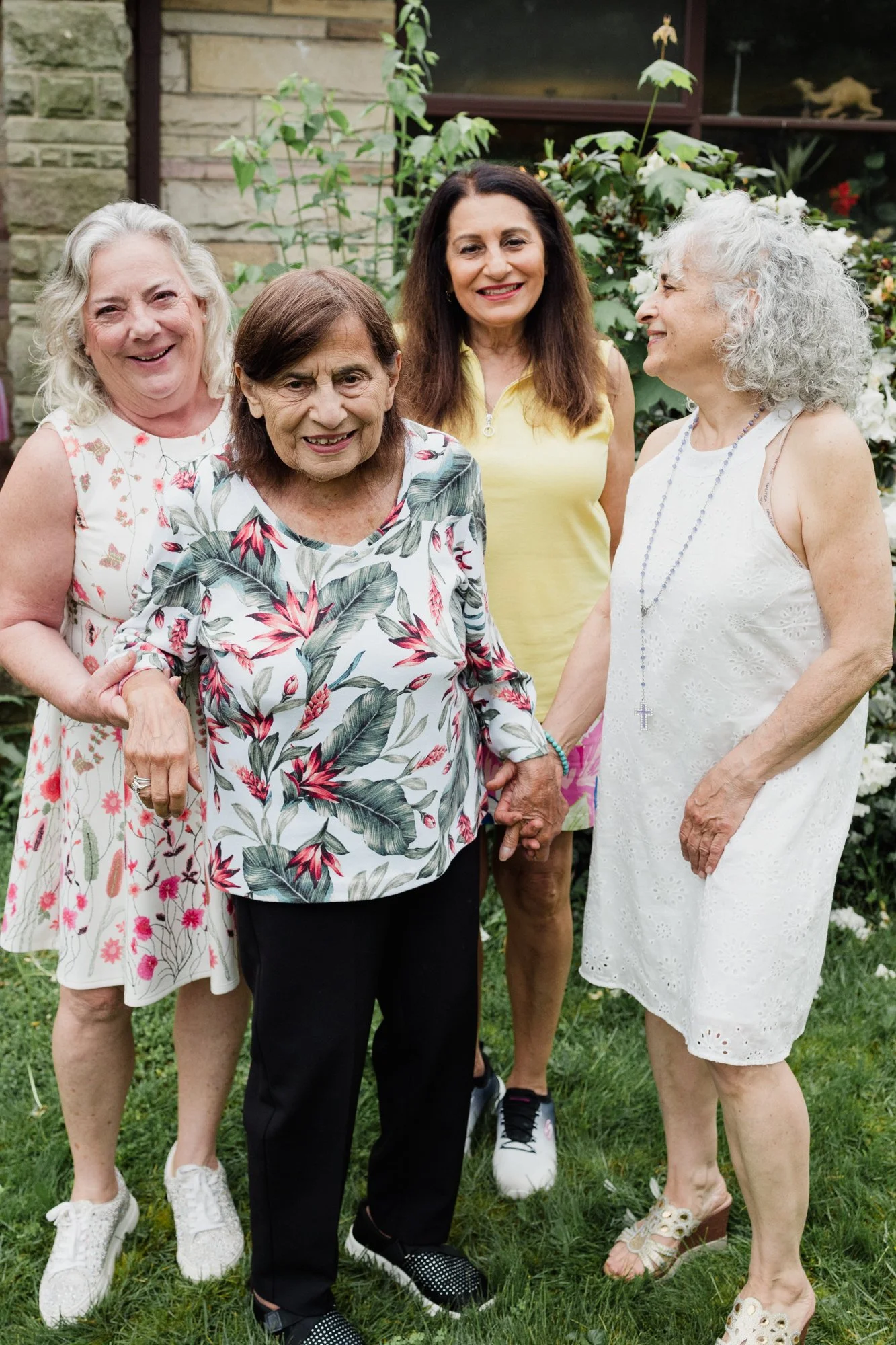 Five women standing together outdoors in a garden, smiling and holding hands.