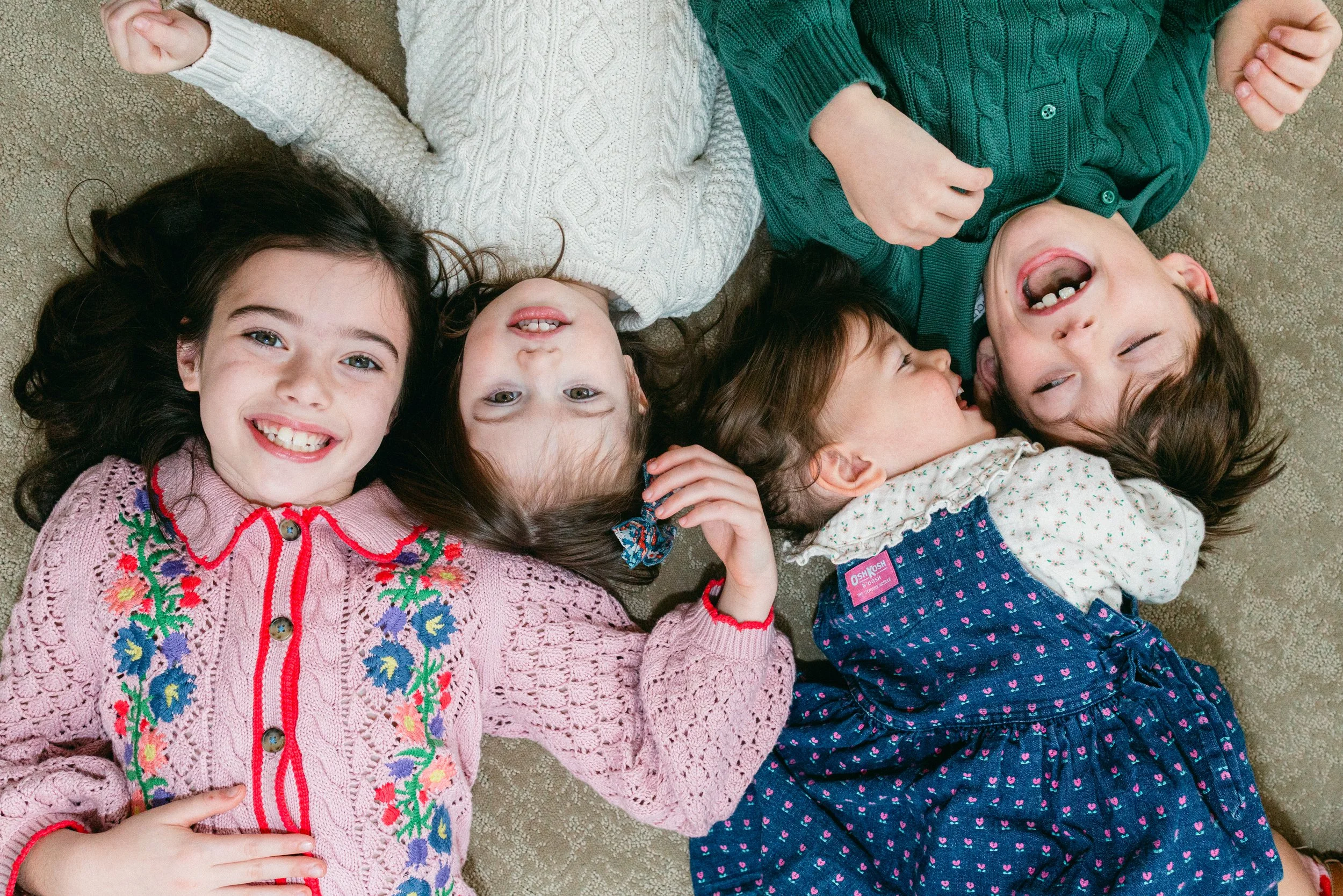 Four young girls lying on a carpeted floor, smiling and laughing, with their heads close together, wearing colorful clothes.