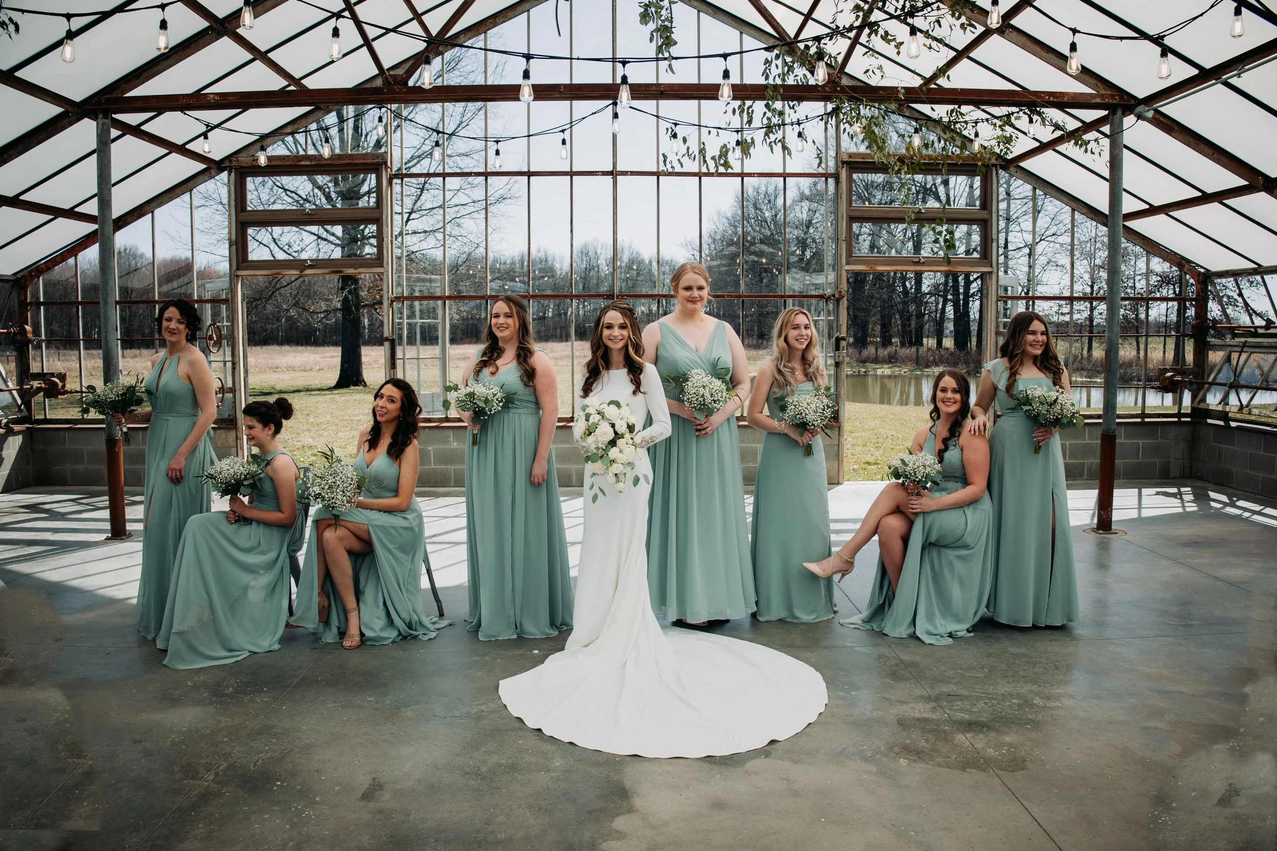 A bride in a white wedding gown holding a bouquet of white flowers stands with eight bridesmaids in mint green dresses, each holding bouquets of white and green flowers, inside a greenhouse with large glass windows and doors showing an outdoor landsc