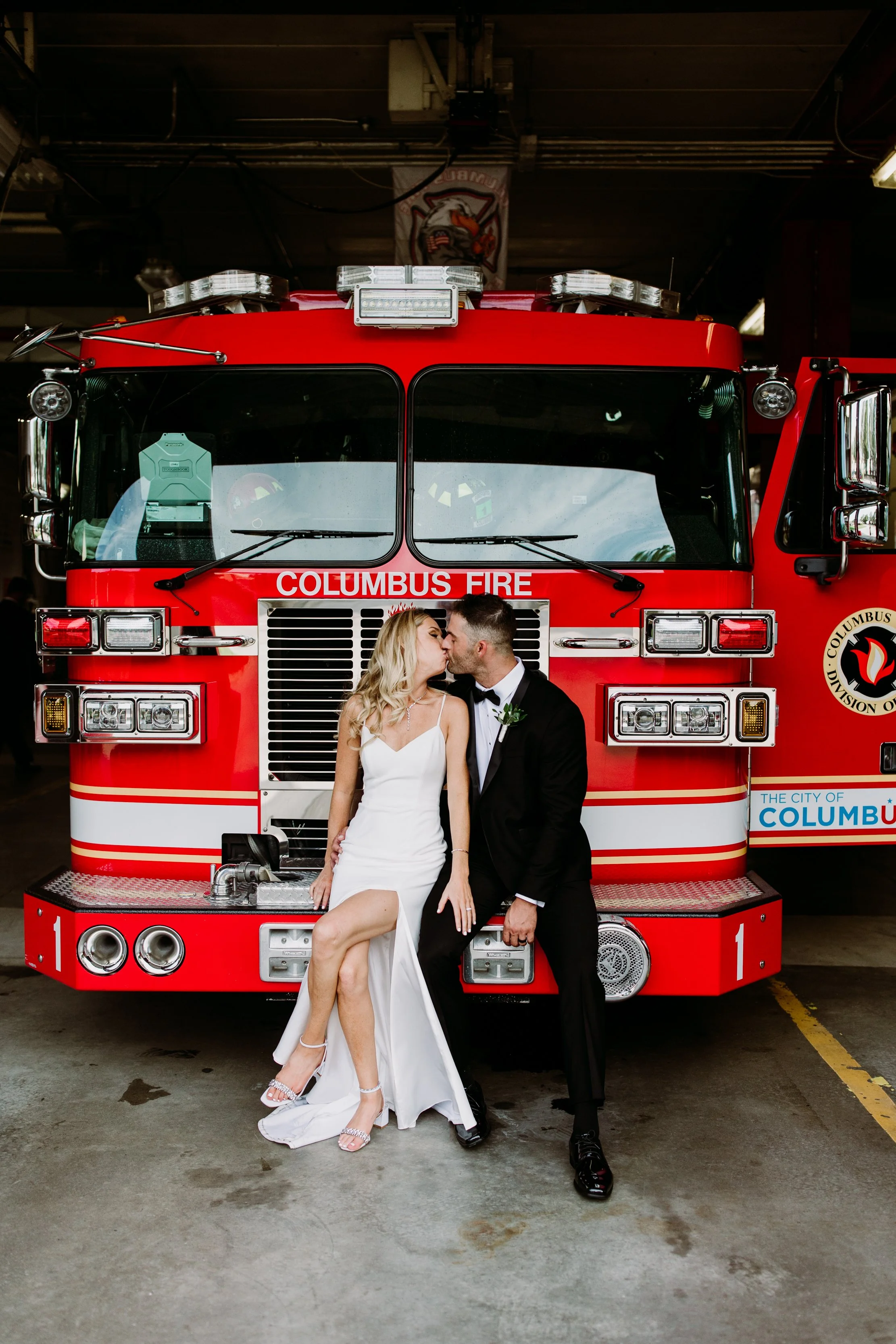 A couple dressed in wedding attire kissing in front of a red Columbus fire truck inside a fire station.