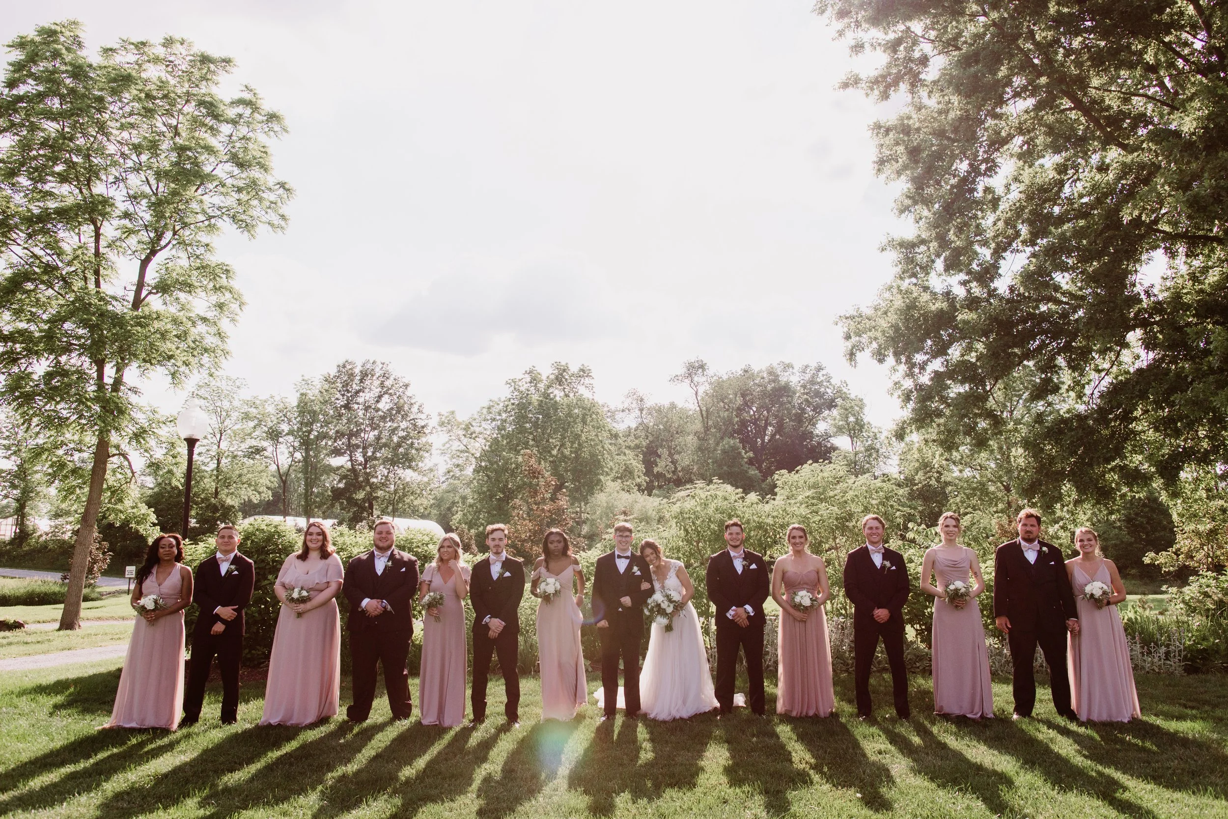 A group of 14 people, including bridesmaids and groomsmen, standing outdoors in a line on a grassy field surrounded by tall trees under a partly cloudy sky, during a wedding celebration.