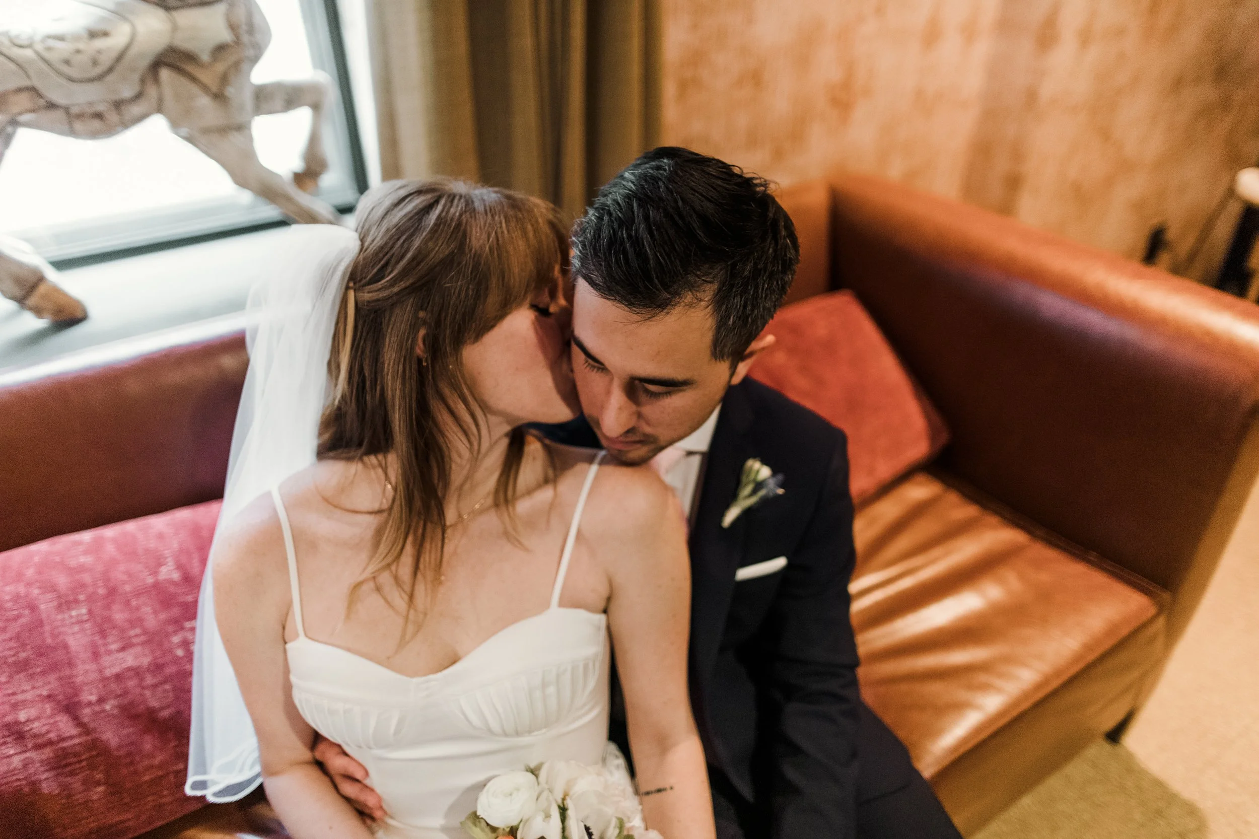 Bride and groom sharing an intimate moment on a leather couch, with the bride kissing the groom's cheek and holding a bouquet of white flowers.
