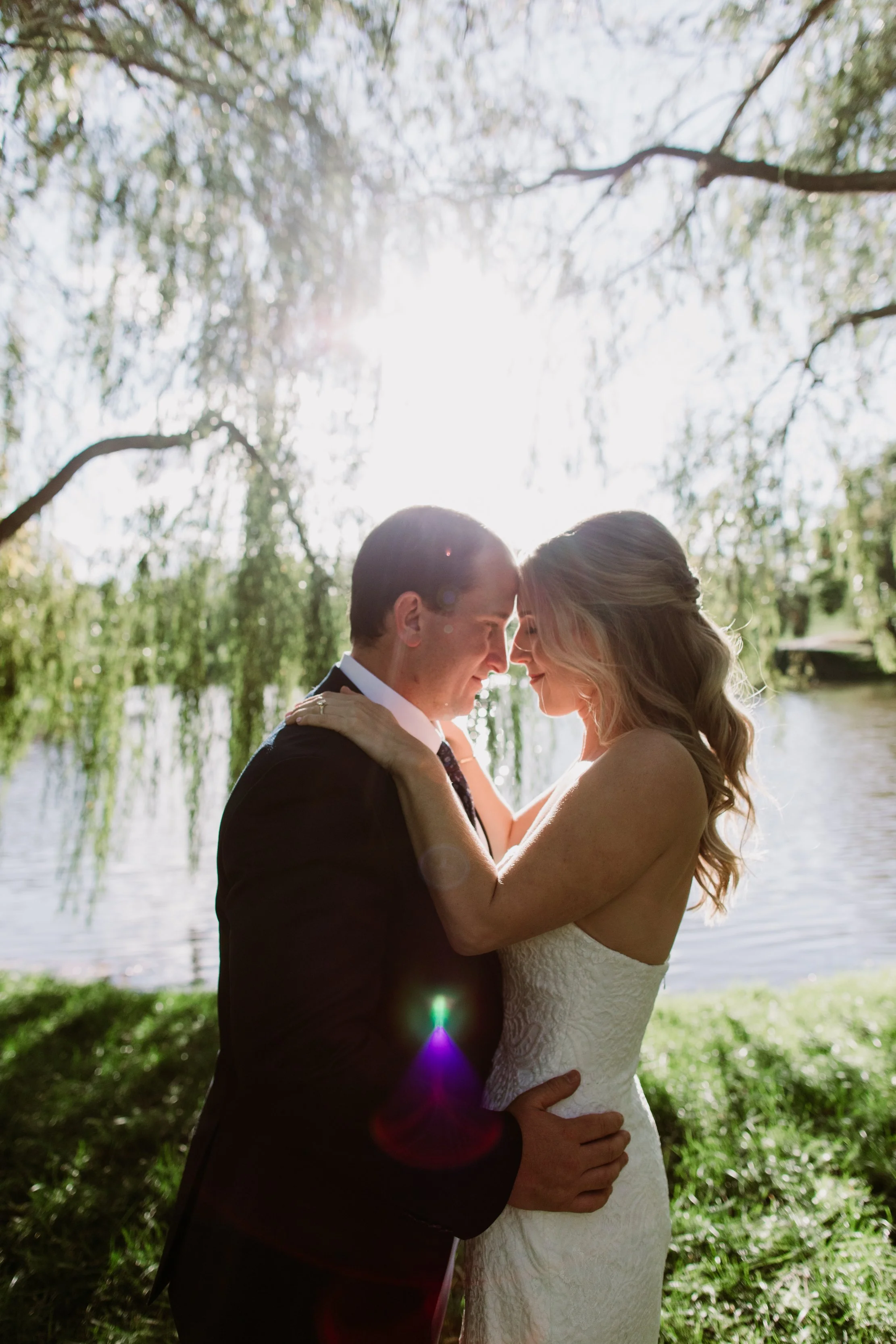 A newlywed couple shares an intimate moment outdoors near a body of water, with the sun shining behind them through tree branches, creating a warm glow and lens flare.