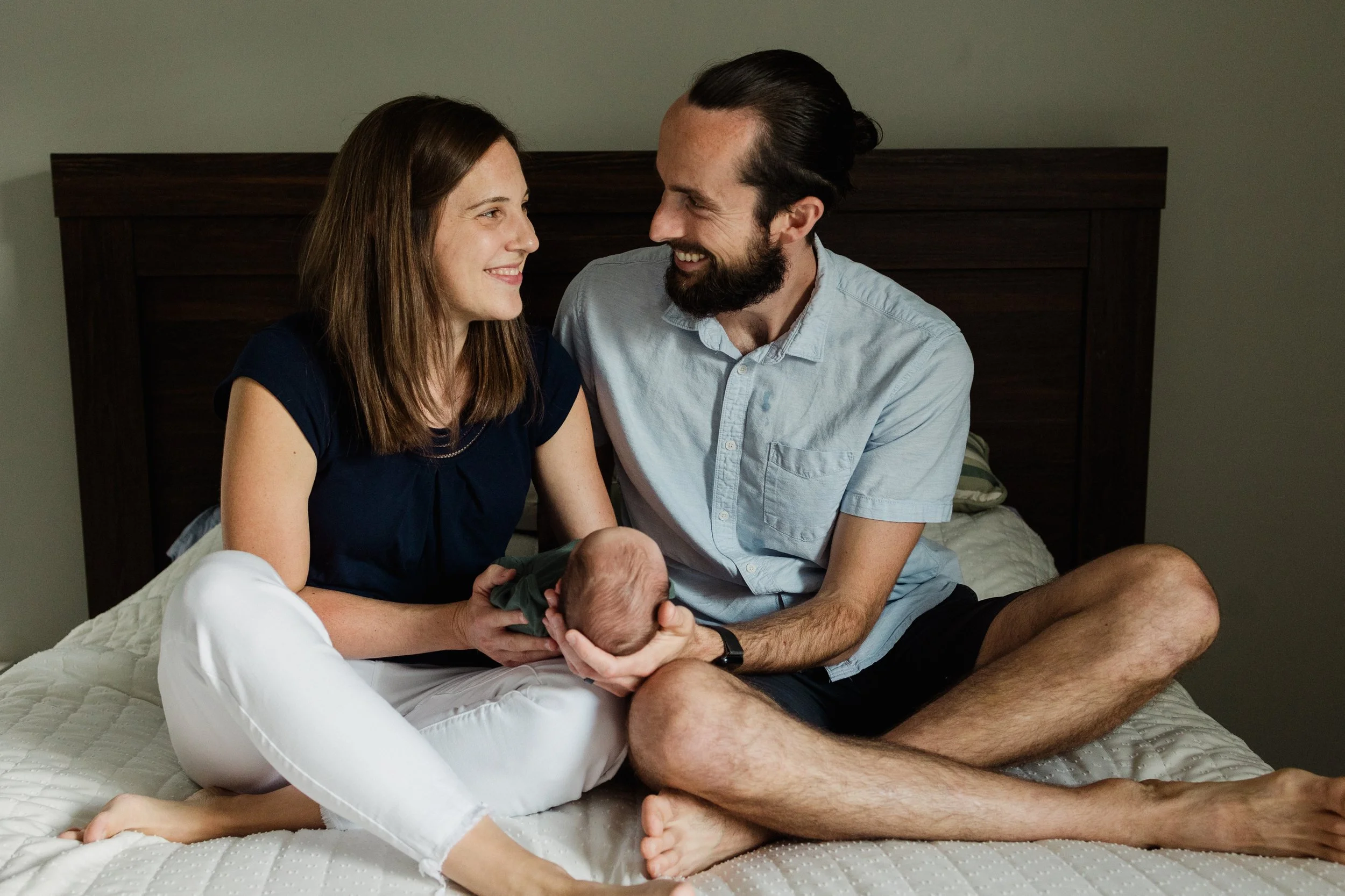 A happy couple sitting on a bed with their newborn baby, smiling at each other.