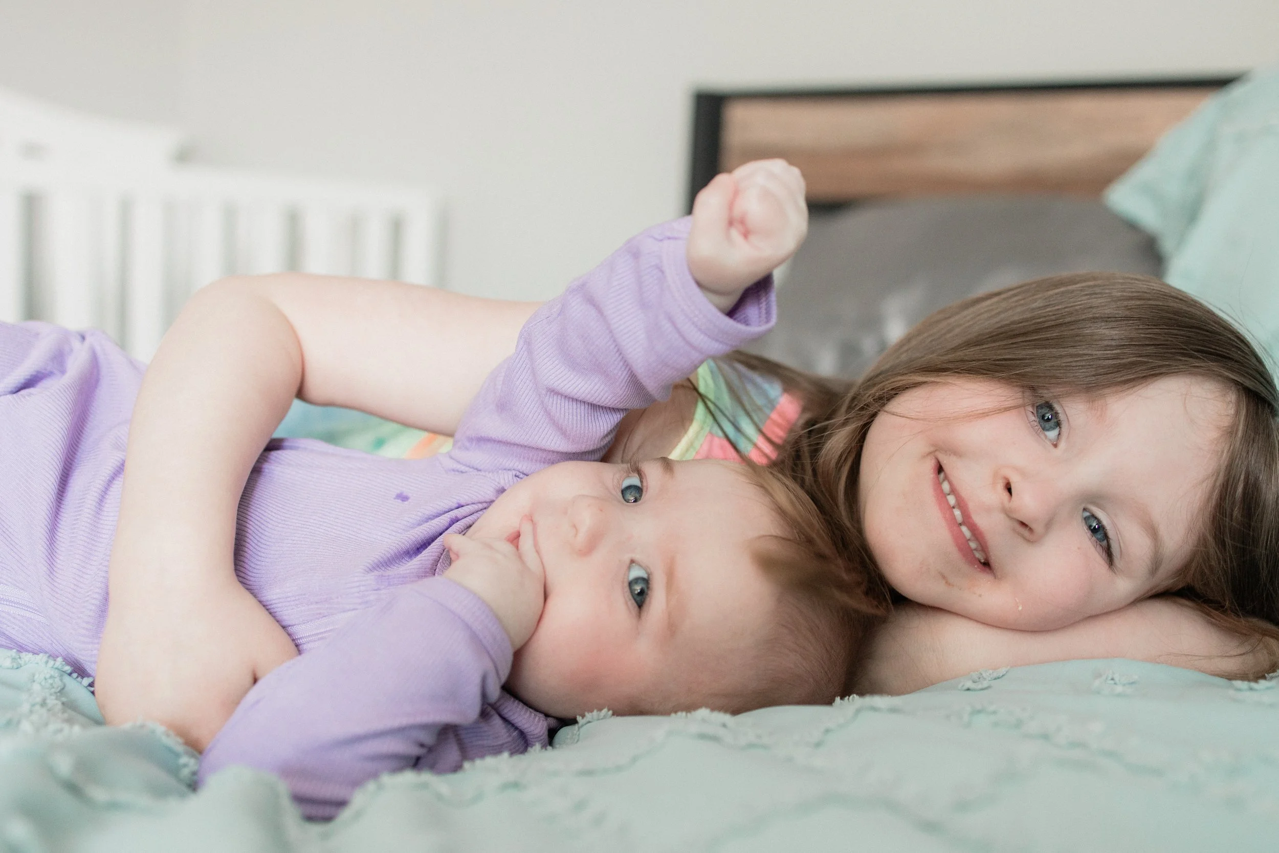 Two young girls lying on a bed, smiling, with the older girl resting her head on her arm and the younger girl with her thumb in her mouth.