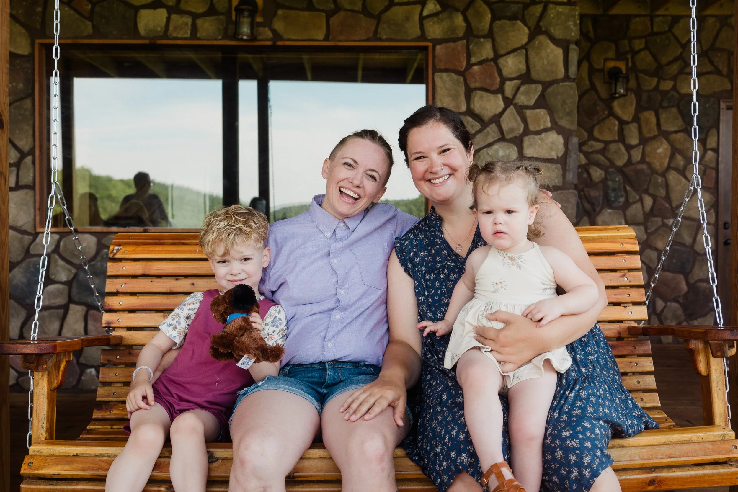 Four people sitting on a wooden swing bench outside, smiling at the camera, with a stone wall and window behind them. Two children, two women, in casual summer clothes.