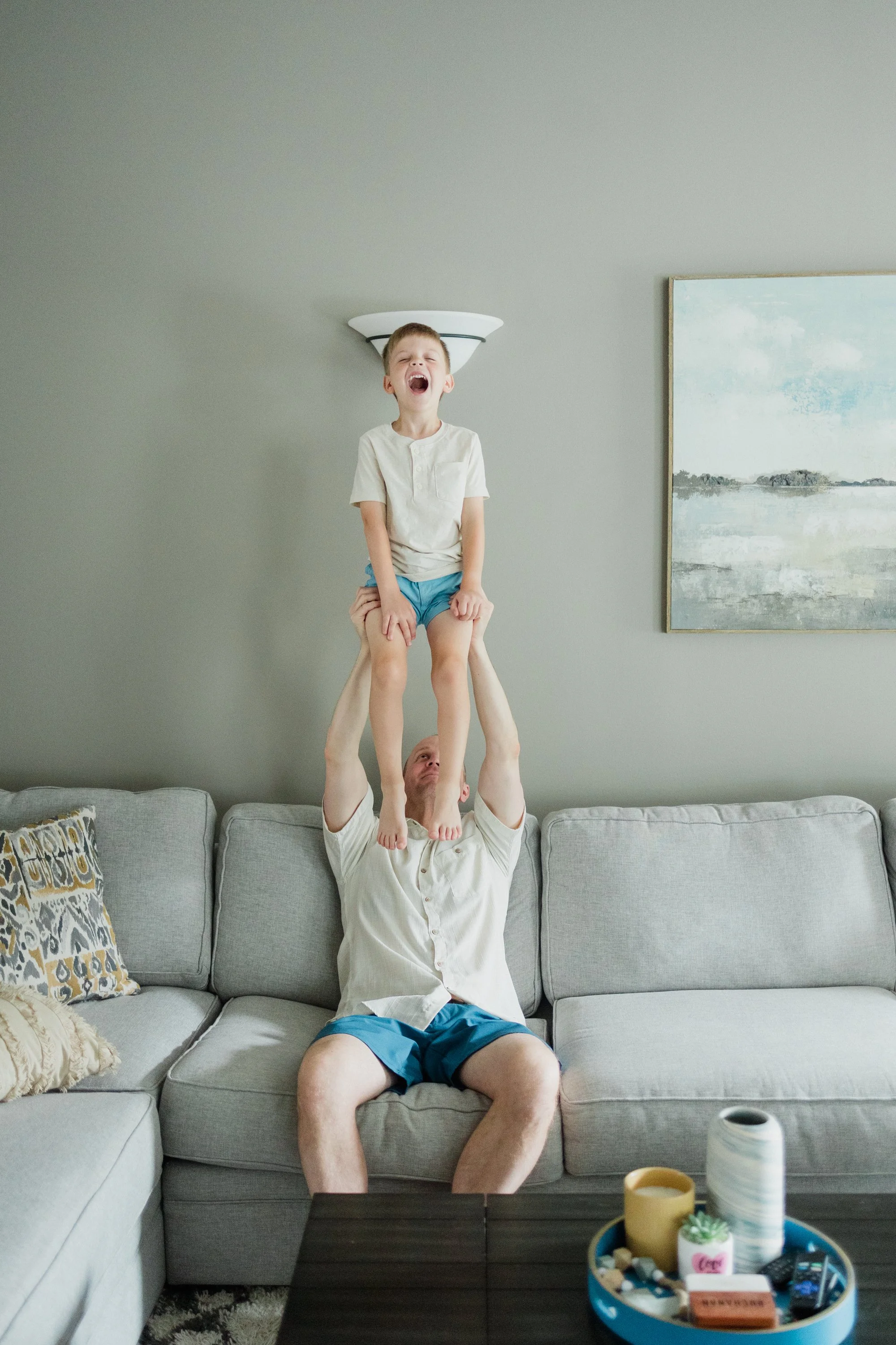 A man sitting on a gray couch lifting a boy above his head. The boy is laughing and wearing a white shirt and blue shorts. A painting is on the wall behind them, and a tray with various items is on a table in front of the couch.