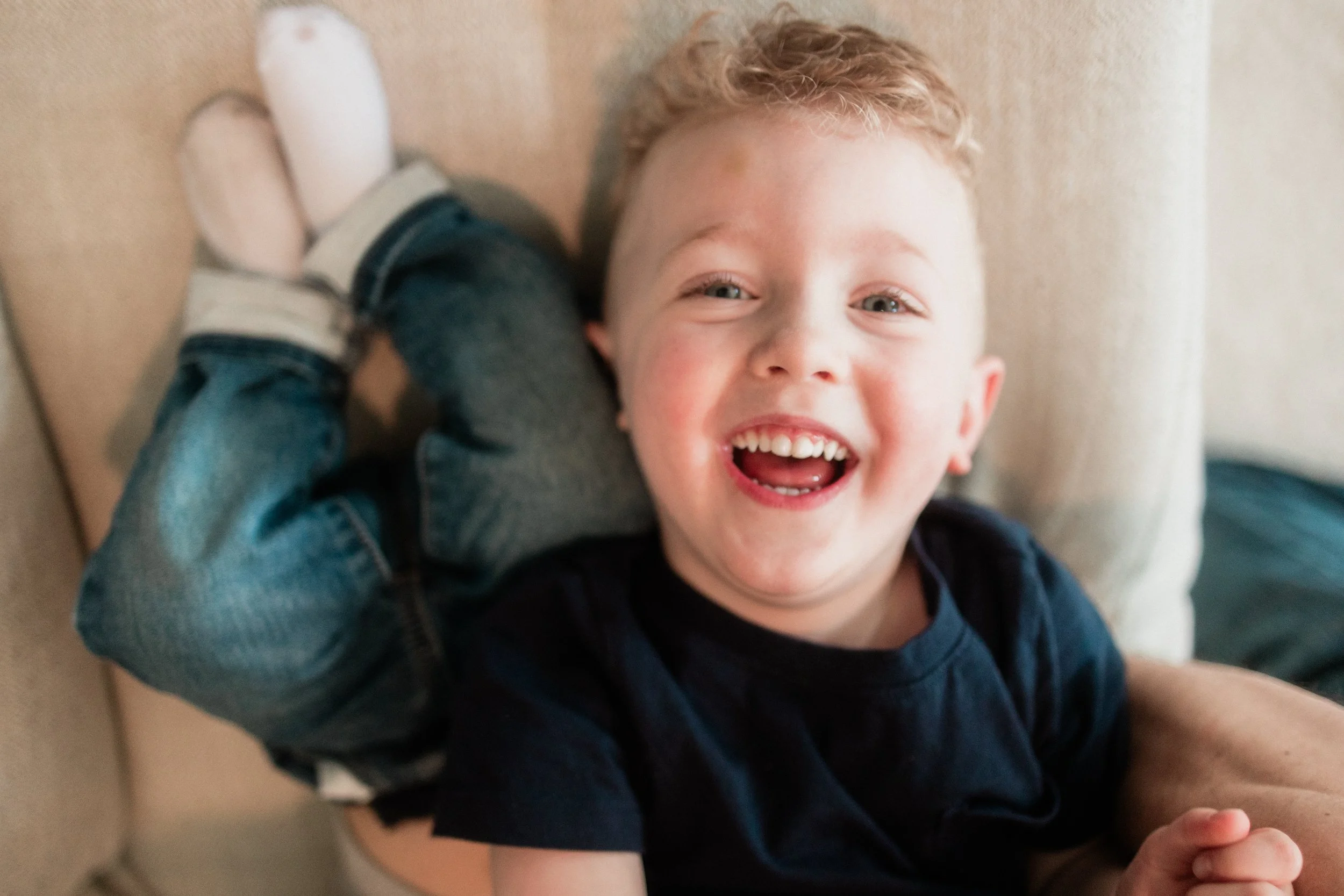 A young boy with curly blond hair, smiling widely, lying on a beige couch, wearing a black t-shirt and denim jeans, with a cushion in the background.