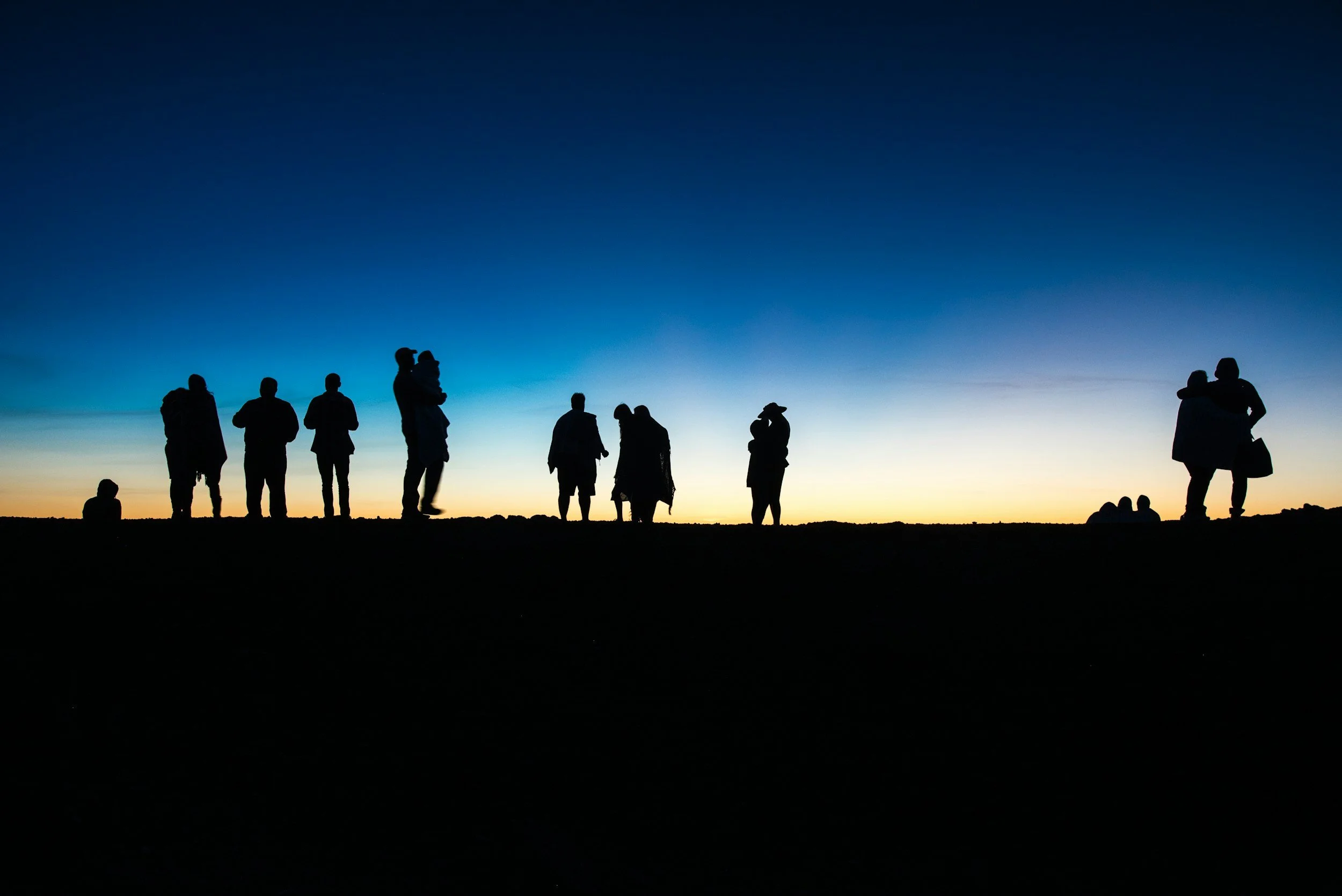 Silhouettes of people standing and walking on a hill during a sunset or sunrise with a colorful sky in the background.