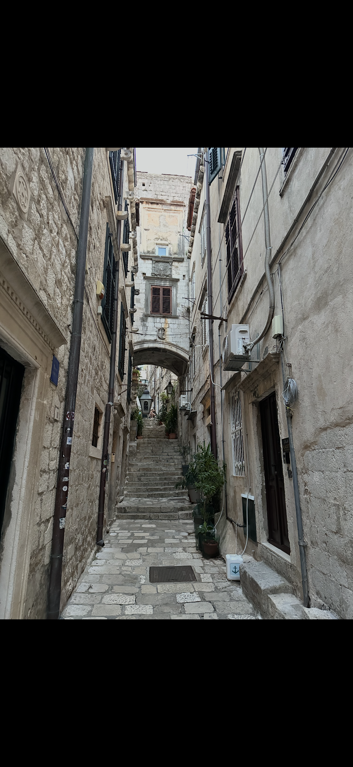 A narrow old European alleyway with stone steps leading up to an archway, surrounded by historic buildings with potted plants along the sides.
