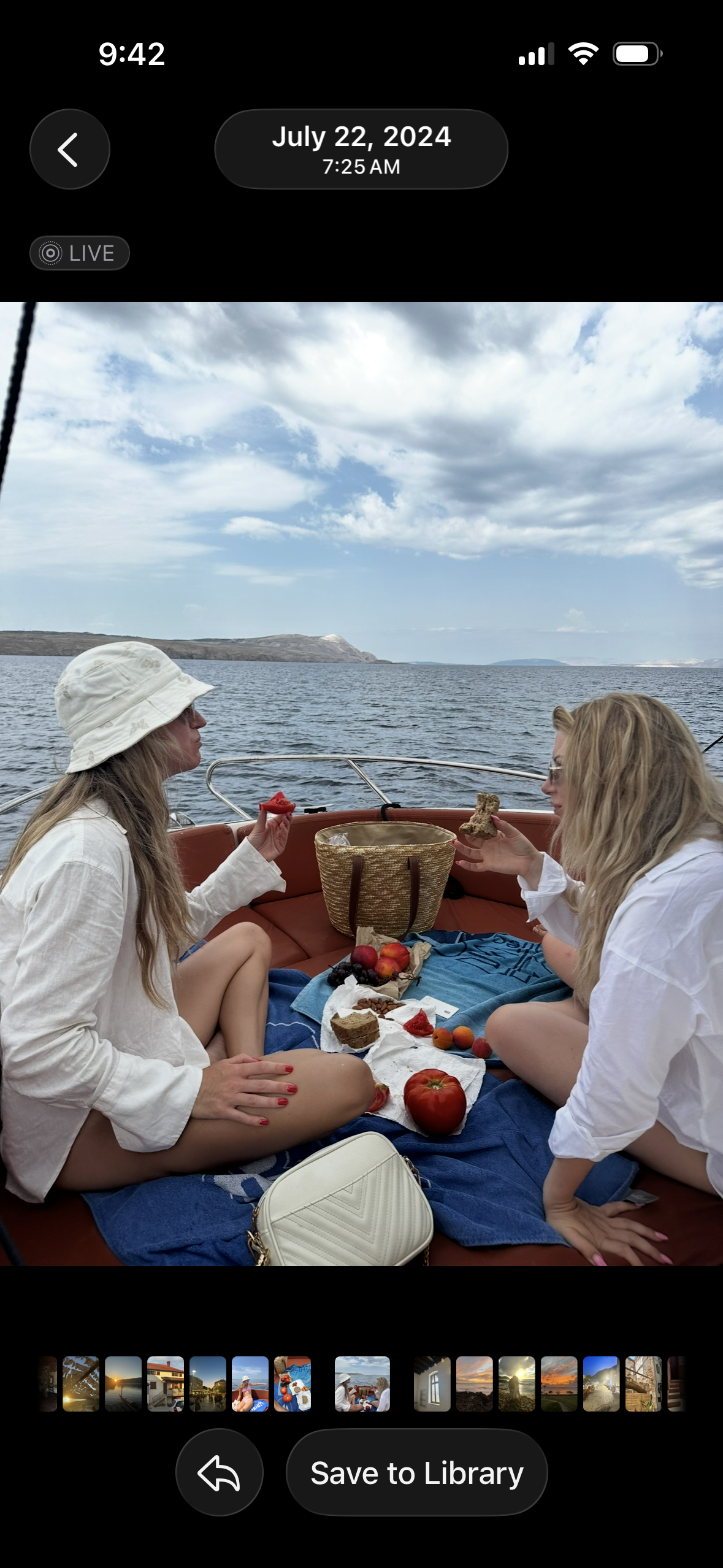 Two women having a picnic on a boat with a lake and hills in the background, eating slices of cake and surrounded by apples and berries, under a partly cloudy sky.