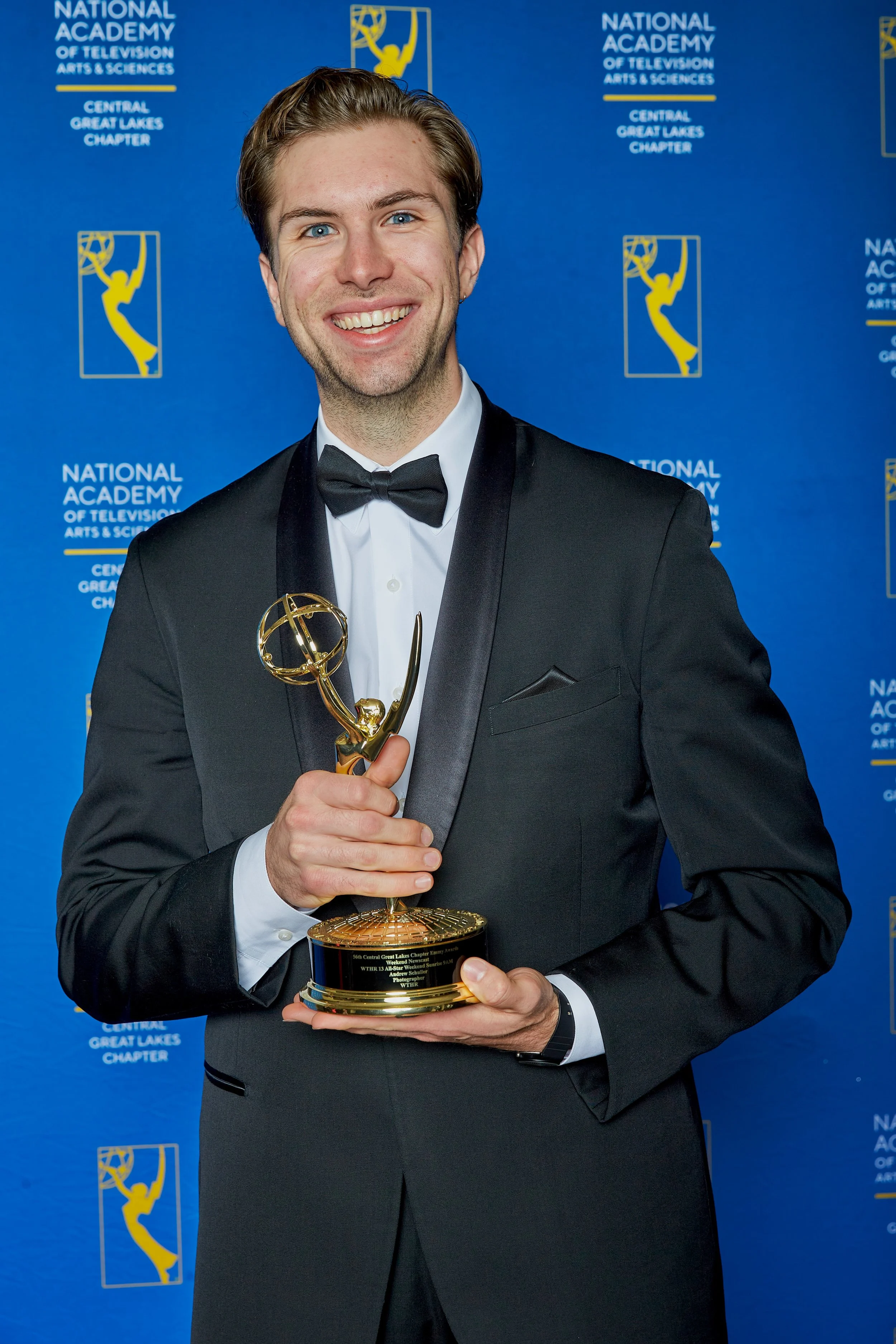 A man wearing a tuxedo and bow tie holding an Emmy award statue, standing in front of a blue backdrop with the logos and text for the National Academy of Television Arts & Sciences, Central Great Lakes Chapter.