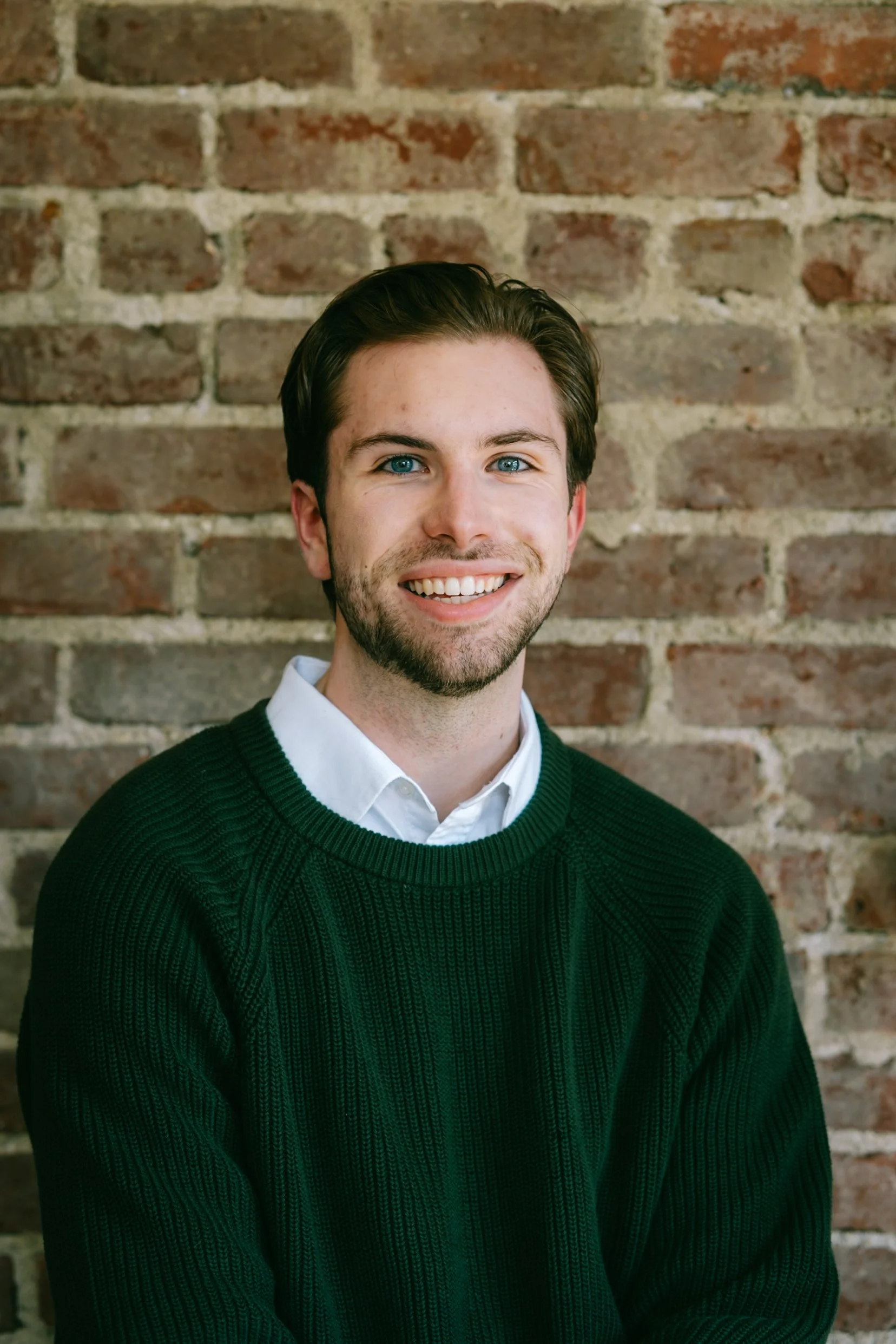 A young man with dark brown hair, blue eyes, and a beard, smiling, wearing a white collared shirt under a dark green knit sweater, standing against a brick wall background.