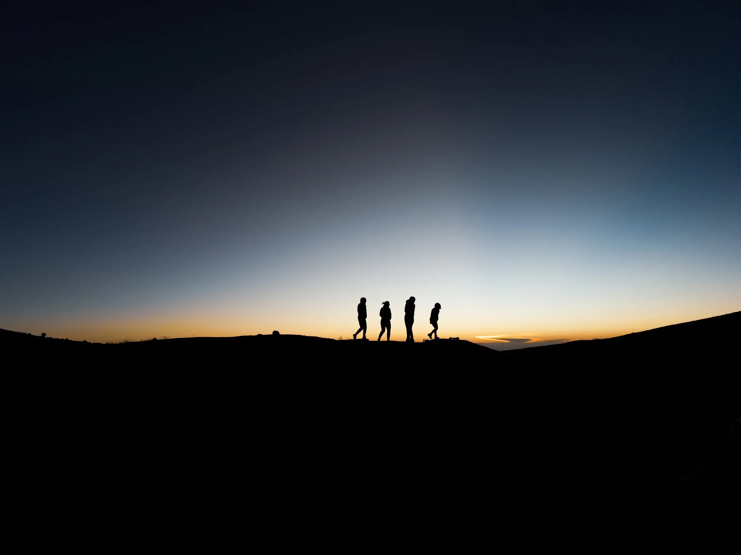 Silhouettes of four people walking on a hill at sunset or sunrise, with a gradient sky transitioning from dark blue to light orange.