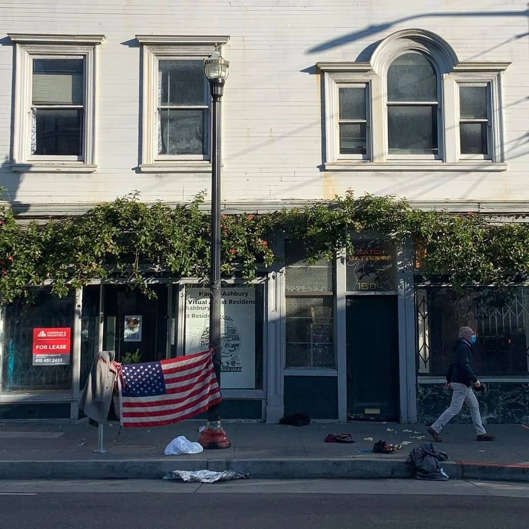 A storefront with an American flag hanging on the sidewalk in front of it, a person wearing a mask walking past, and a street lantern in front of a white building with three windows and decorative trim.