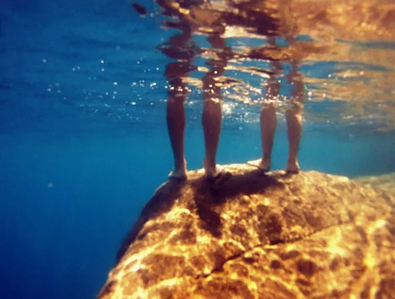 Three people standing on a large submerged rock underwater, with only their legs visible.