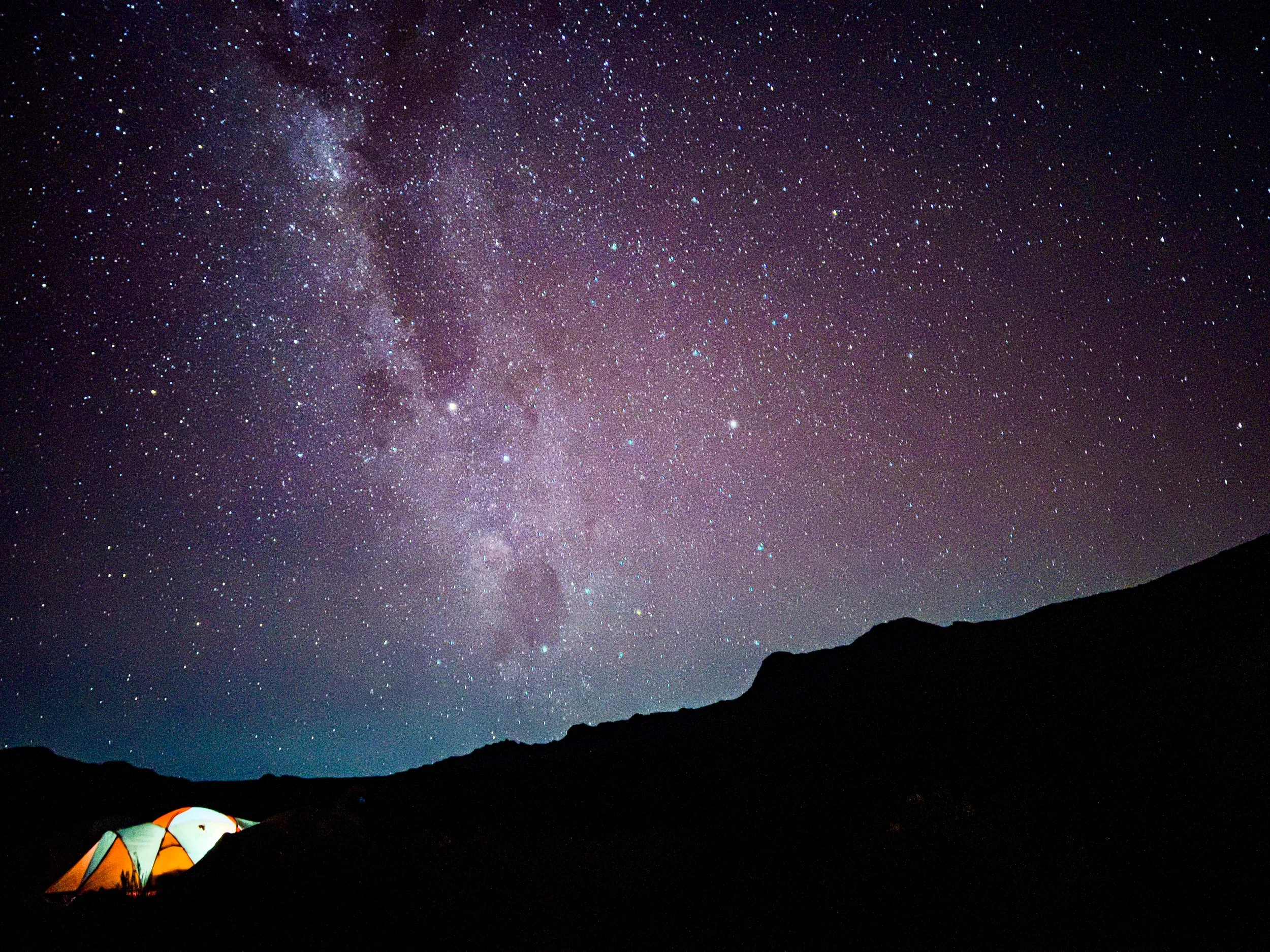A night sky filled with stars and the Milky Way galaxy visible, with a silhouetted mountain range and a brightly lit tent in the foreground.