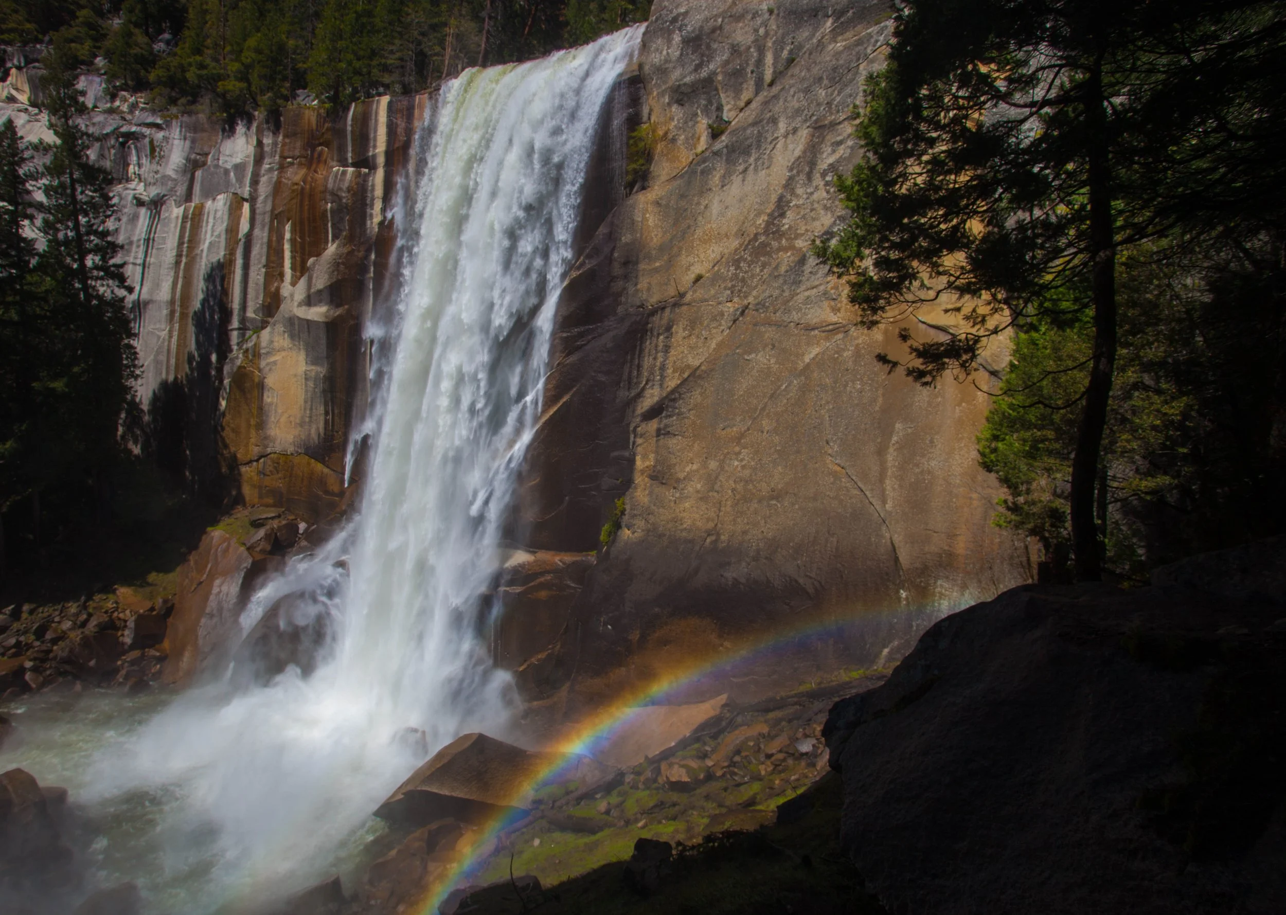 Tall waterfall cascading over rocks and cliffs with a rainbow visible at the base, surrounded by a forest with tall evergreen trees.