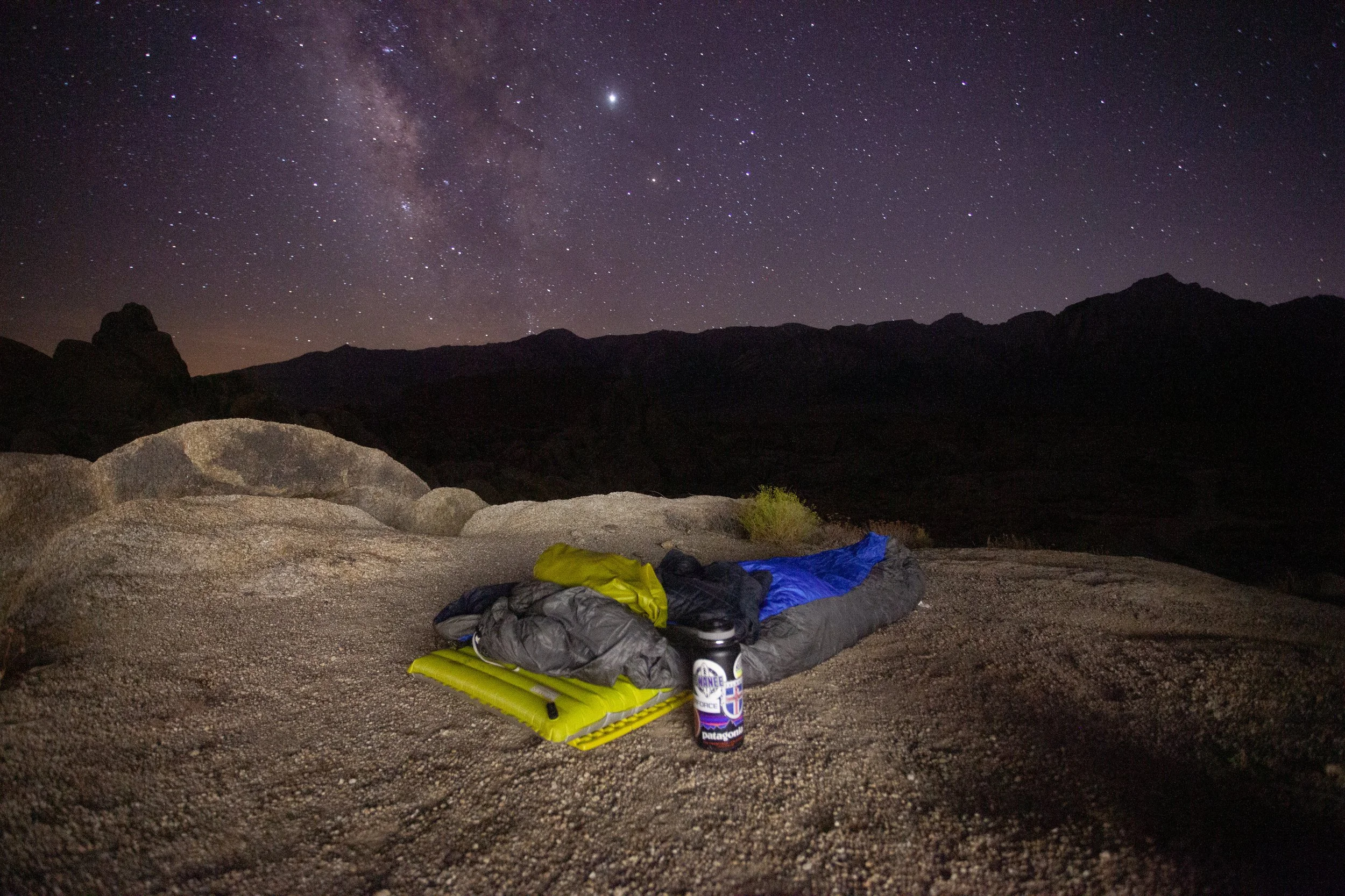 Camping gear with a sleeping bag, mat, and water bottle on rocky terrain under a starry night sky with the Milky Way.