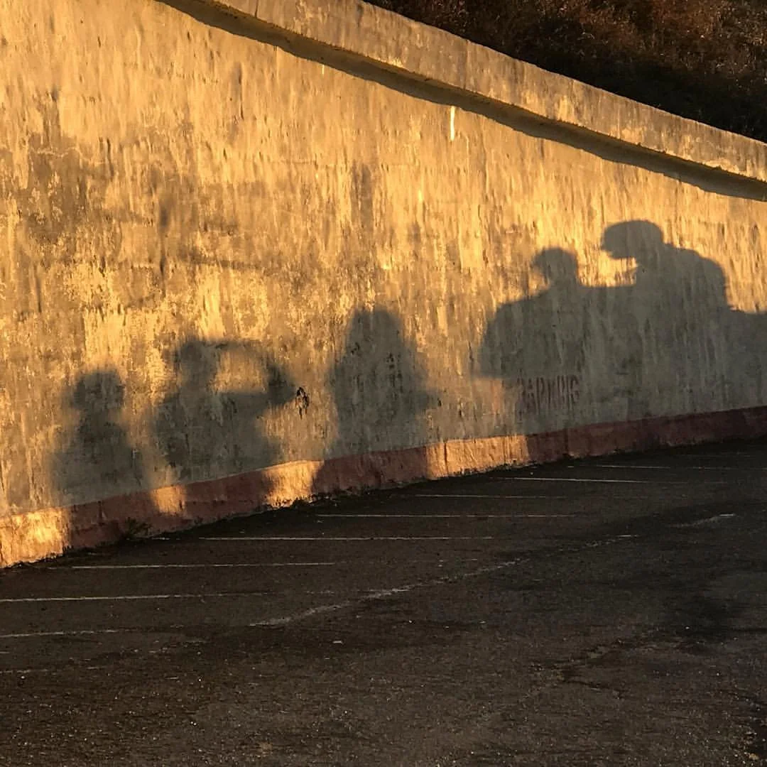 Shadows of three people cast on a weathered concrete wall in a parking lot during sunset.