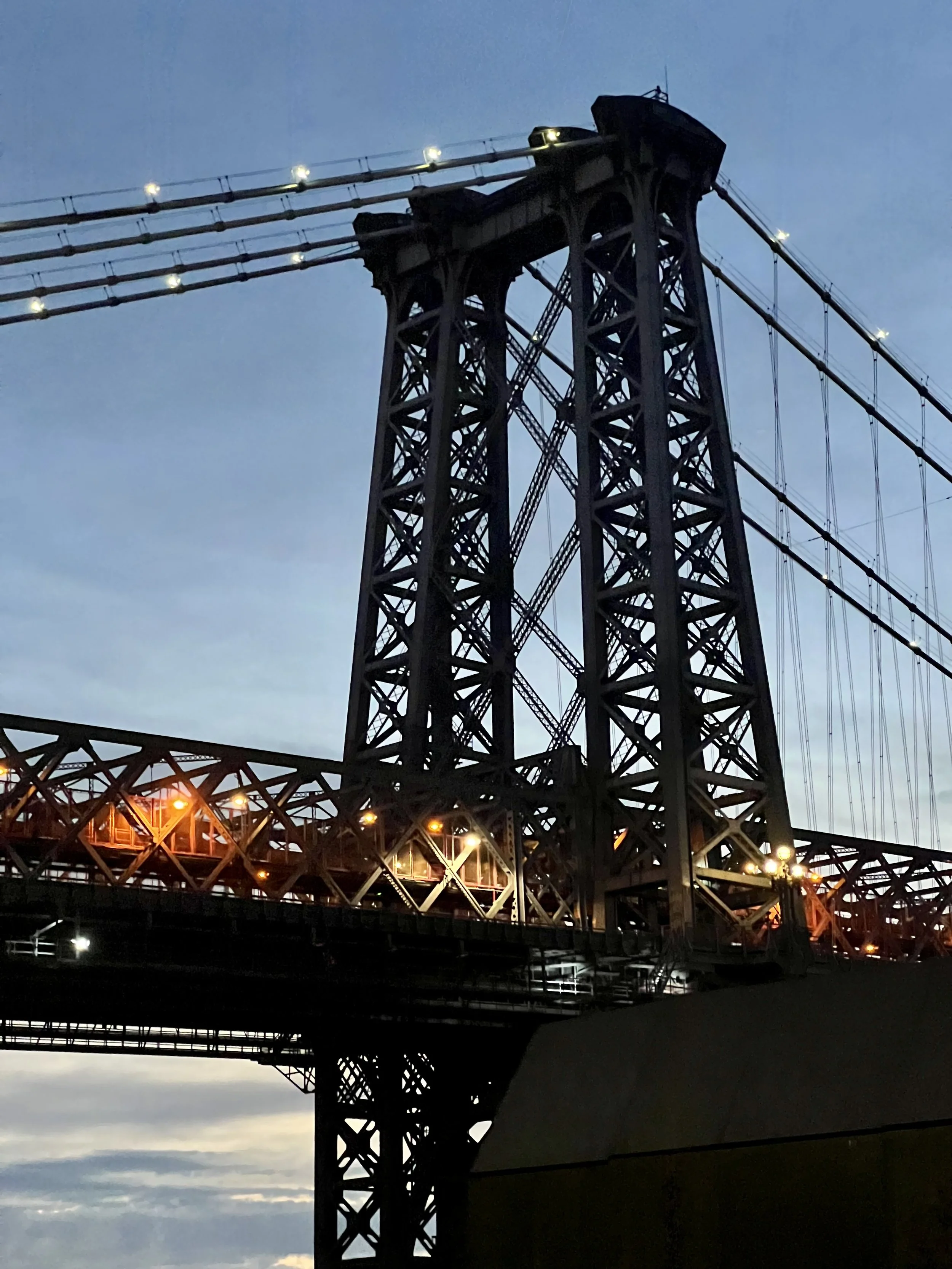 Close-up of a bridge tower with cables at dusk, illuminated by streetlights against a darkening sky.