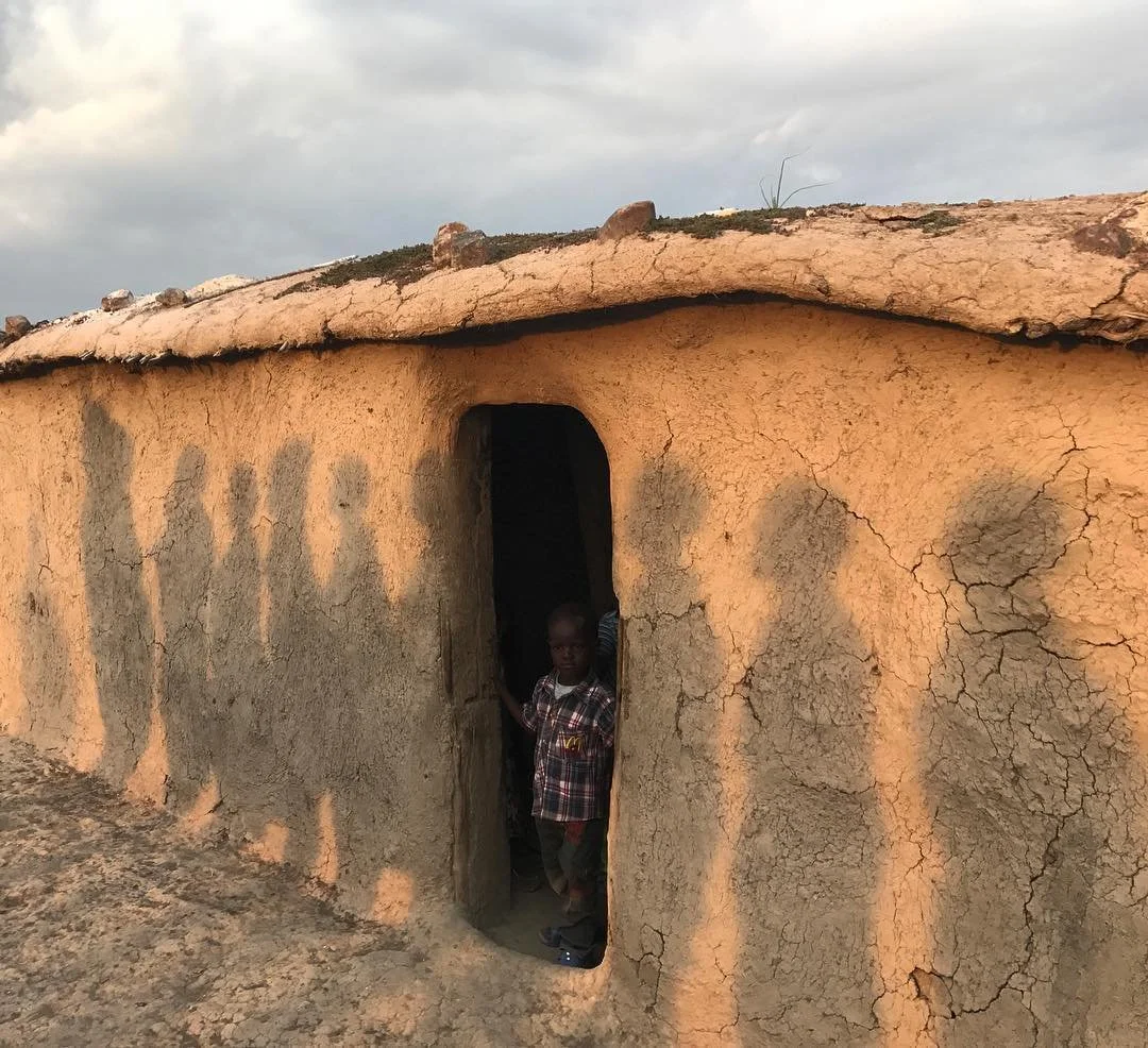 Child standing in the doorway of a mud house with a cracked, earthy exterior, under a cloudy sky.