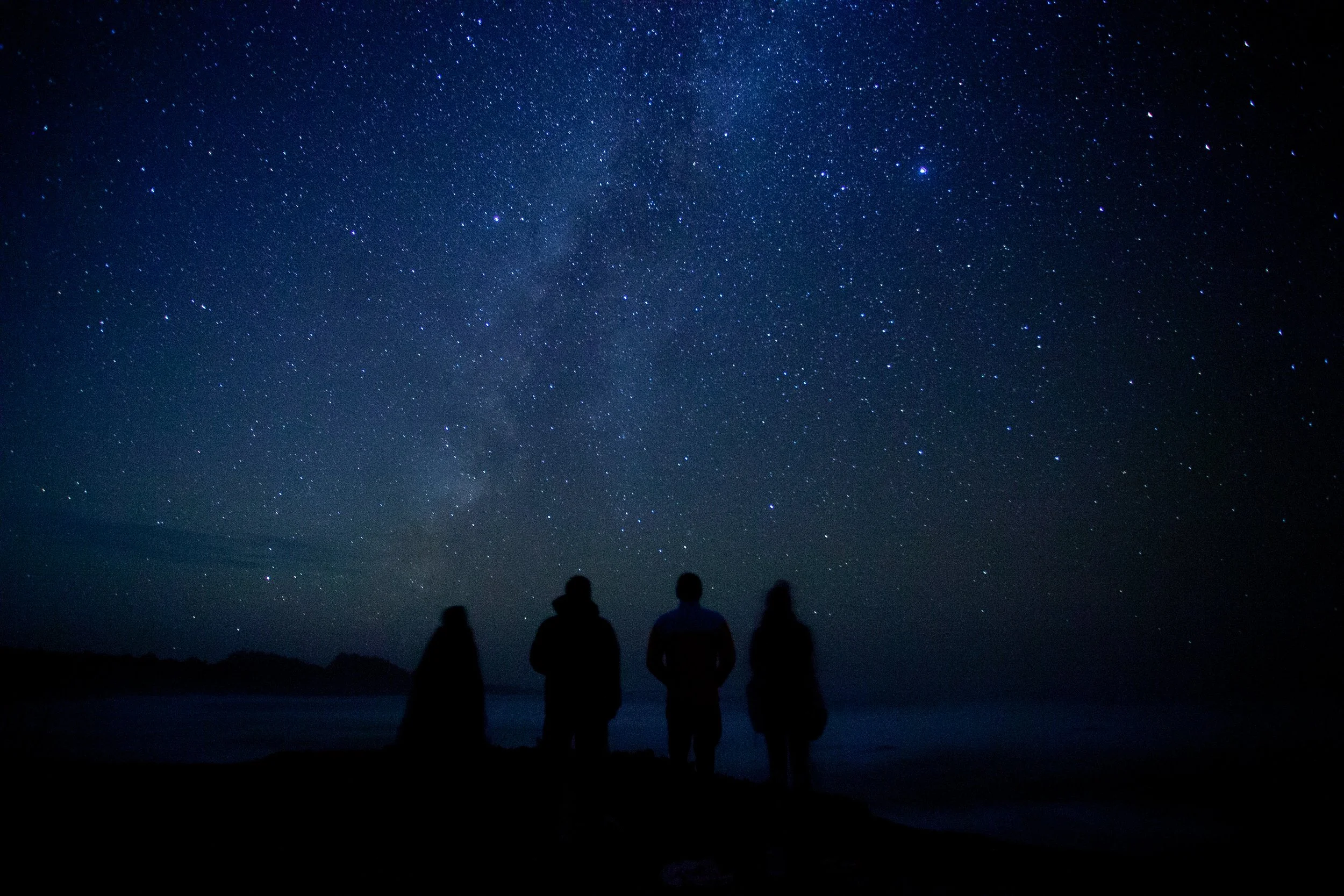 Four people standing on a dark beach at night, staring up at a star-filled sky with the Milky Way visible.