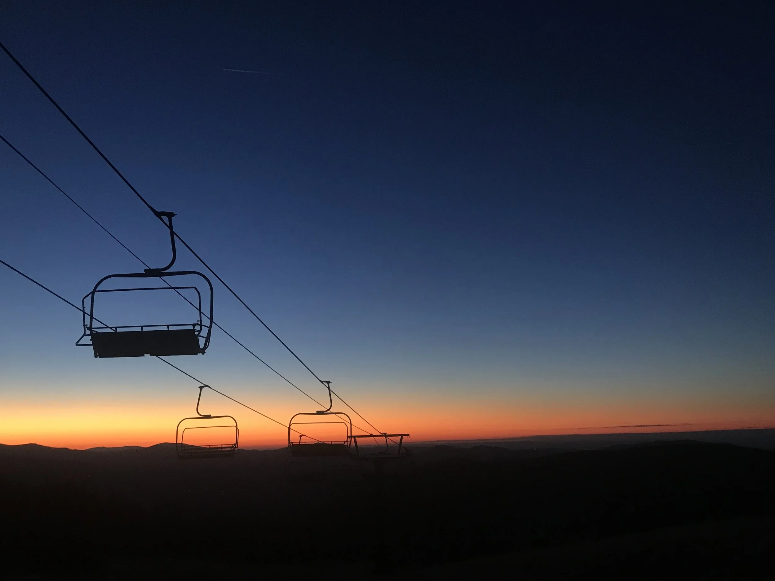 Silhouette of ski lift chairs against a sunset sky with a gradient from orange to deep blue.