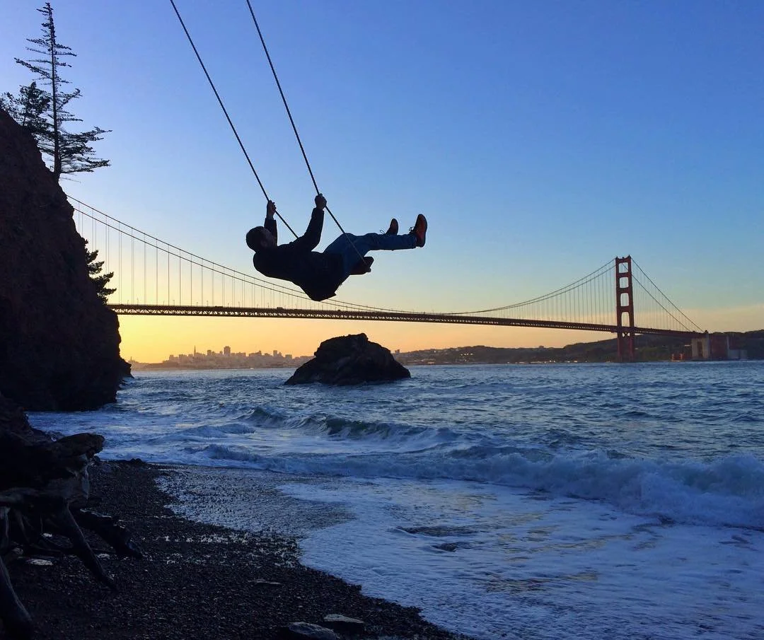 Person swinging on a ropes swing at the beach with the Golden Gate Bridge and city skyline in the background during sunset.