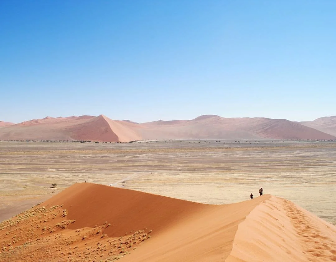 Sand dunes in a desert with two people walking on a small dune, distant mountains under a clear blue sky.