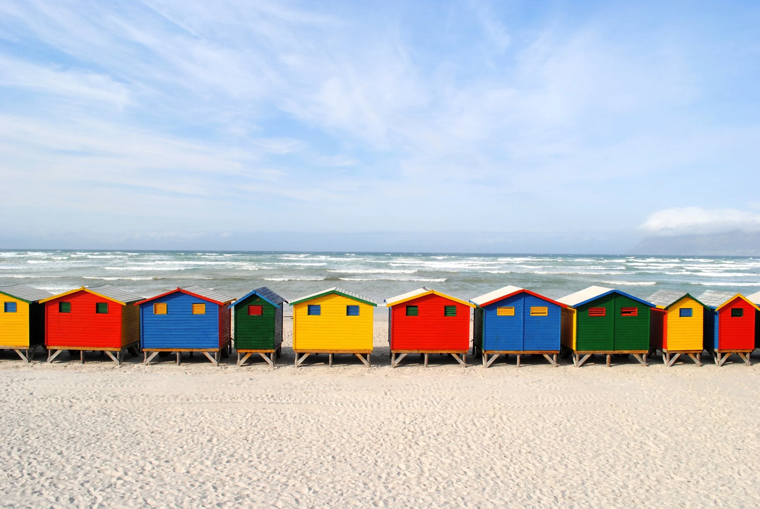 Colorful beach huts on sand with ocean waves and blue sky in the background.
