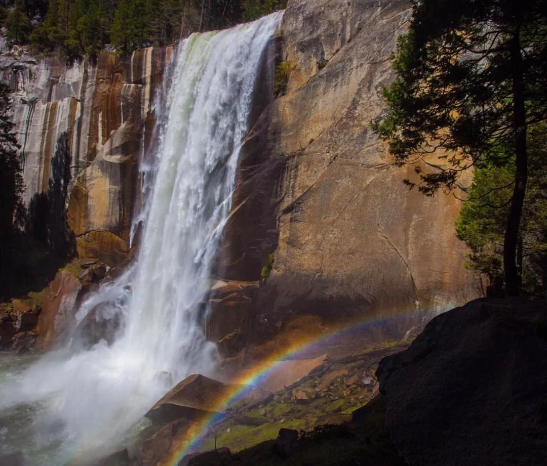A tall waterfall cascading down a rocky cliff with a rainbow at its base and lush green trees surrounding the scene.