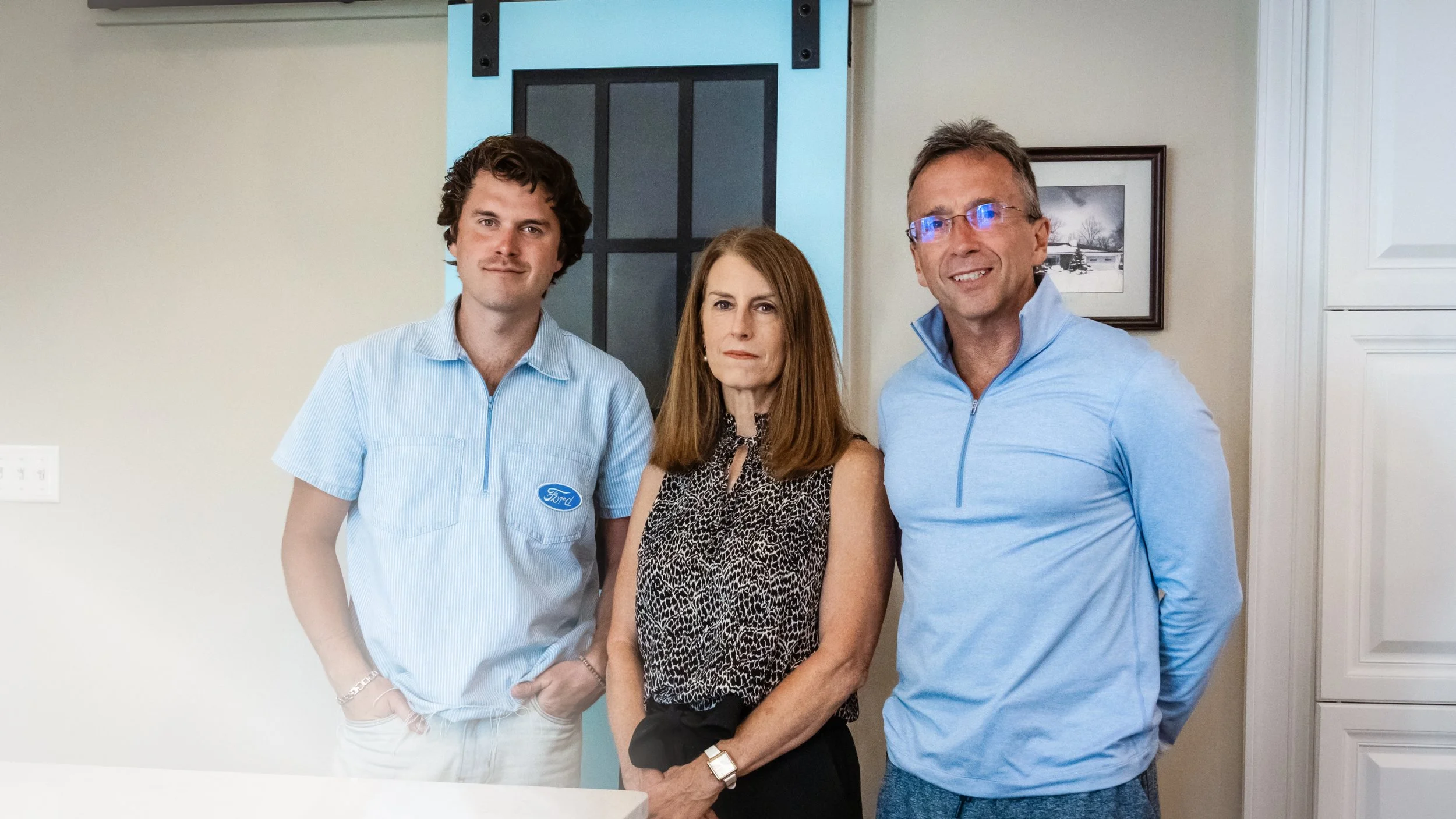 Three people standing indoors in front of a light-colored wall with a picture frame and a blue door.