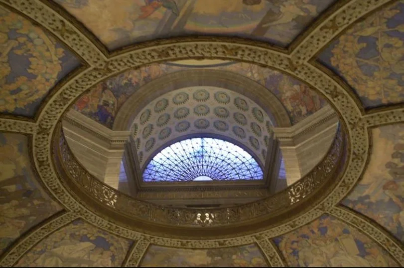 View of an ornate, circular interior ceiling with intricate artwork and a stained glass skylight at the center, framed by decorated arches and a railing.