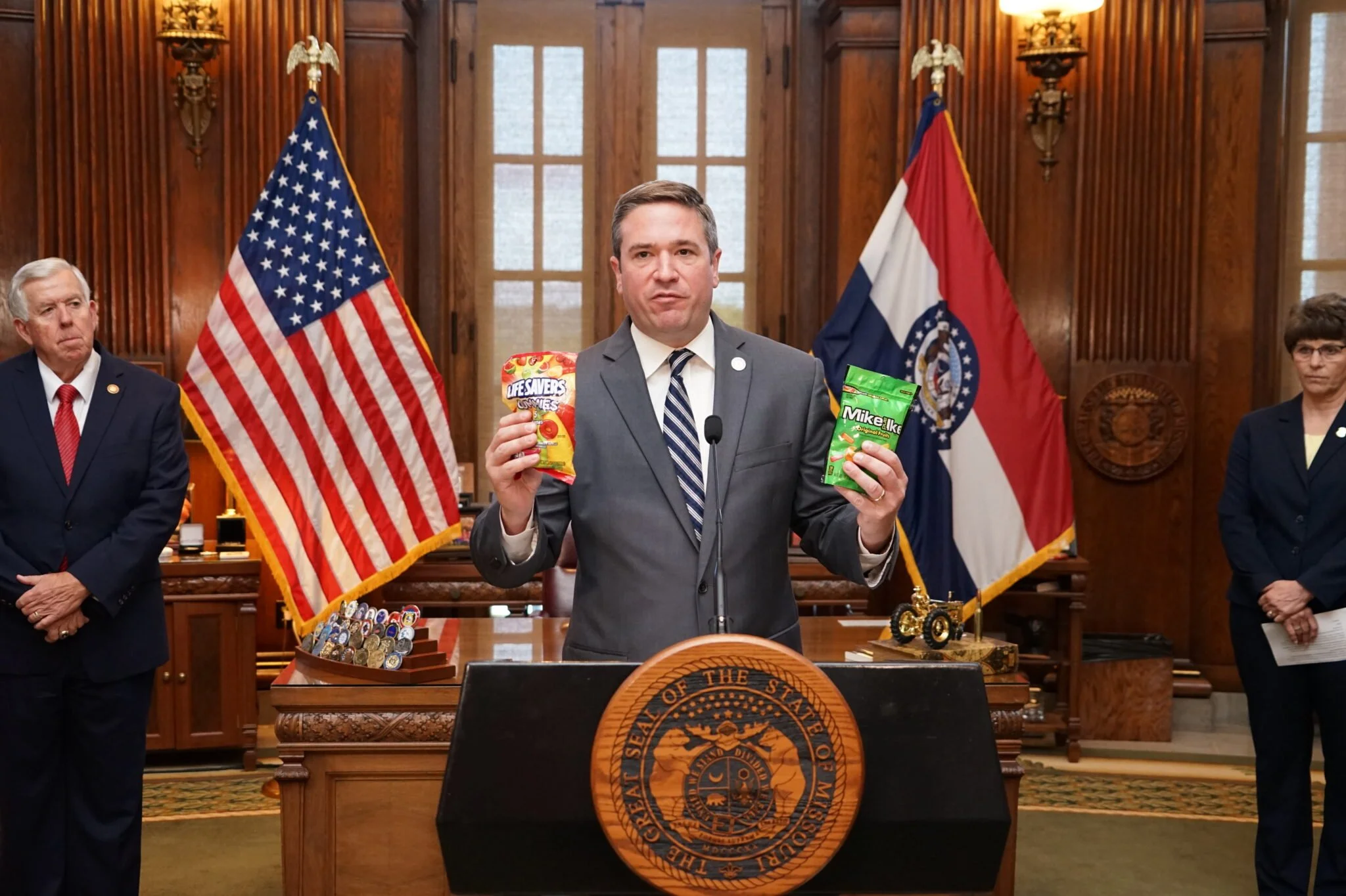 A man in a suit holding bags of snacks at a press conference with flags and officials in the background.
