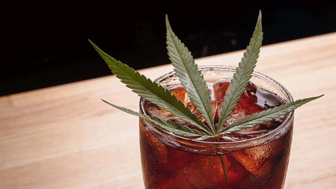 A glass of dark soda with ice and a cannabis leaf as a garnish.