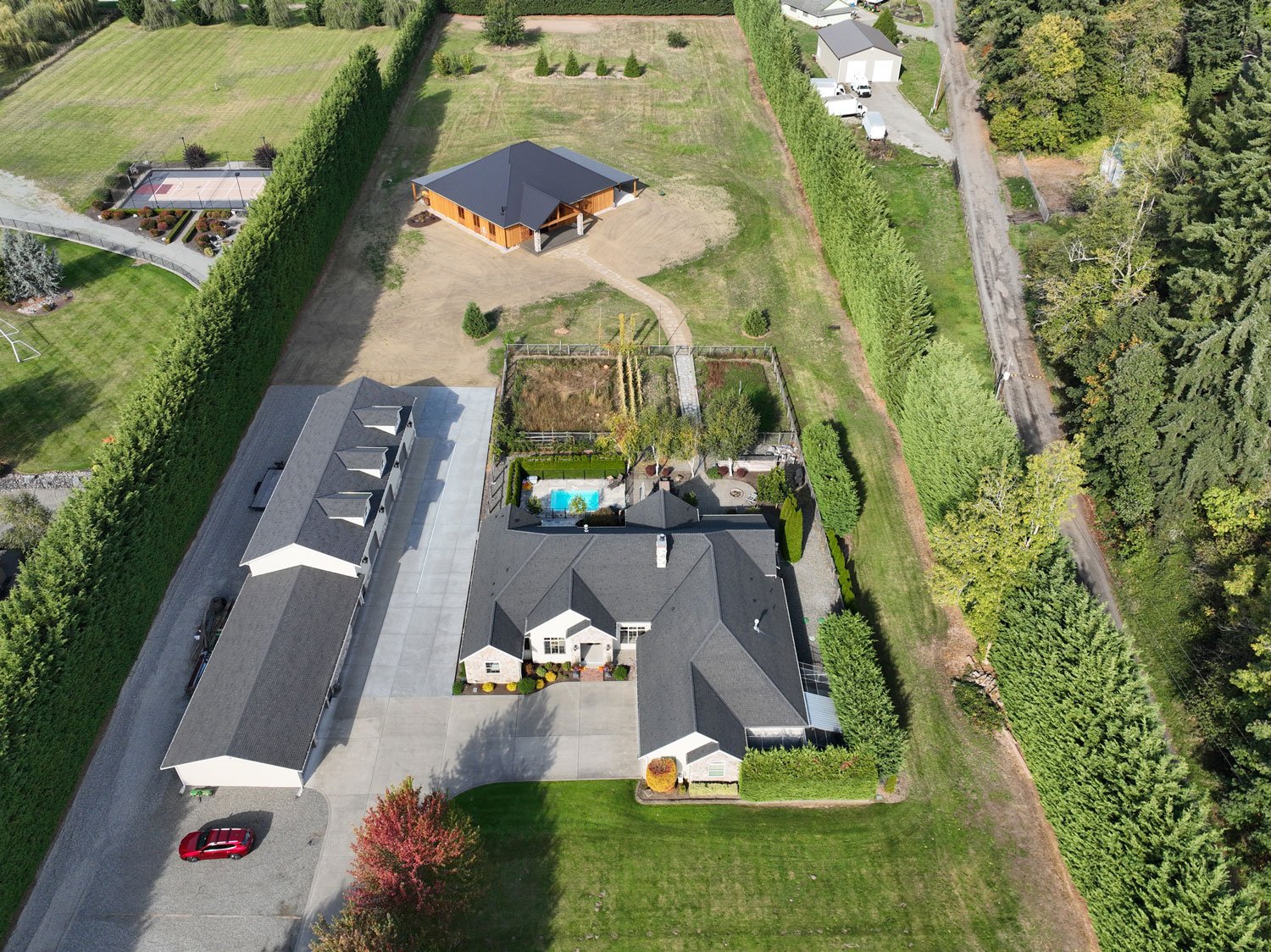 Aerial view of a residential property with a main house, a swimming pool, a large backyard, a detached garage or workshop, and sports court, surrounded by a tall hedge and neighboring areas with trees and grassy fields.