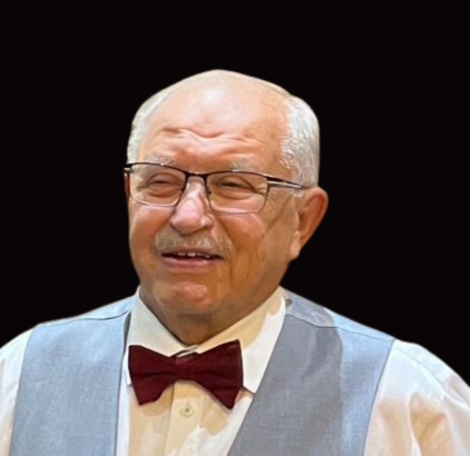 An elderly man with glasses, dressed in a light blue vest, white shirt, and red bow tie, smiling against a black background.
