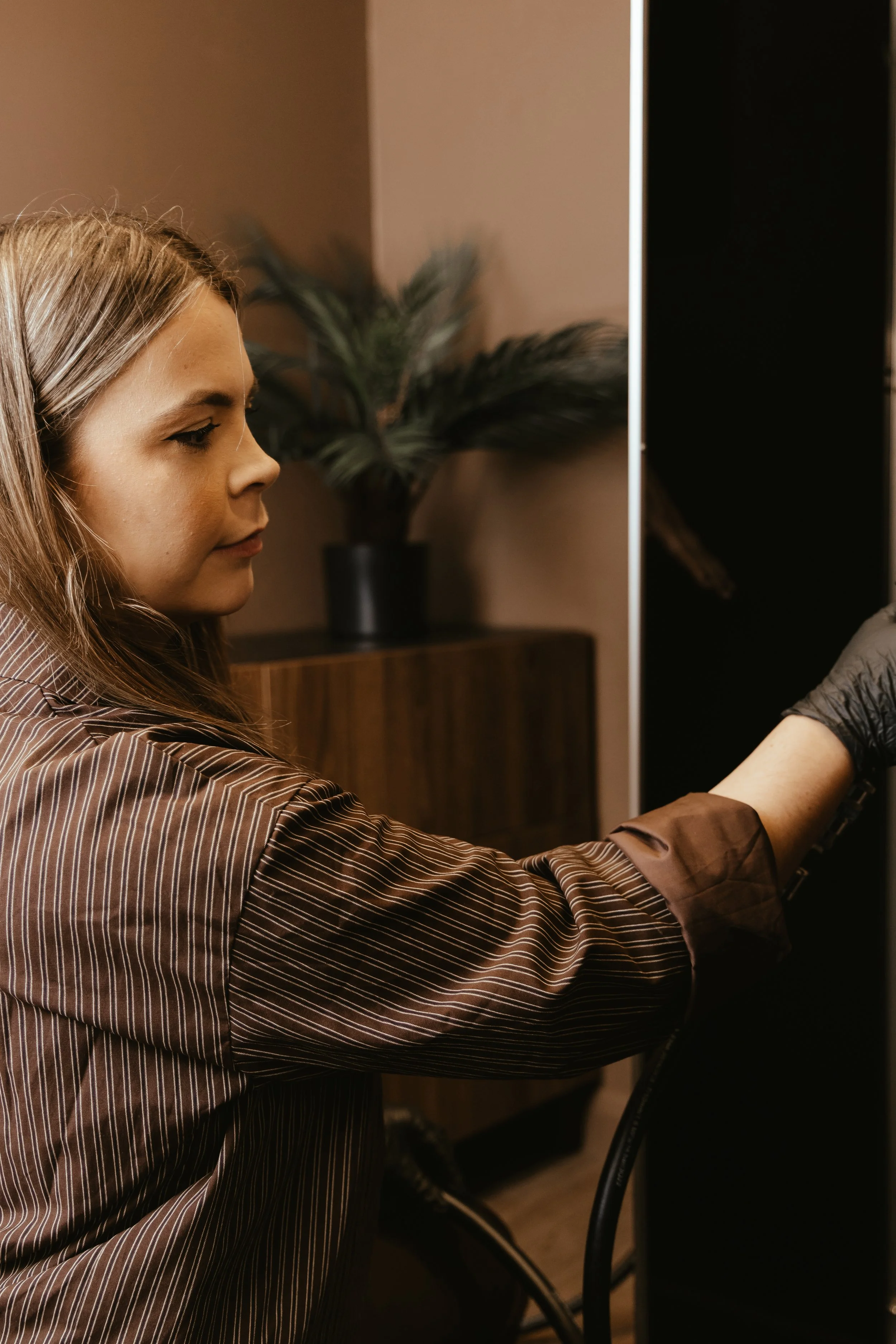 A woman with long hair is plugging in or unplugging an electronic device in a room with a potted plant on a cabinet in the background.