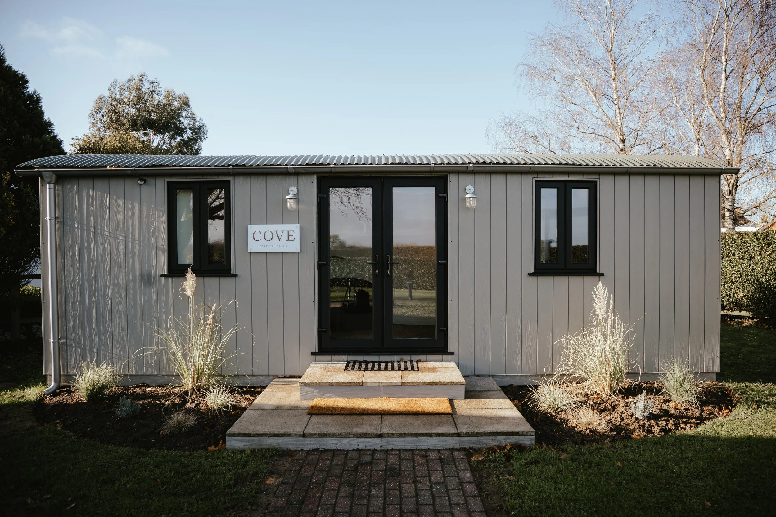 A modern gray tiny house with black-framed windows and a glass door, featuring a sign that reads 'COVE'. The entrance has a small wooden stoop with two steps and a doormat, surrounded by landscaped plants and a brick pathway leading up to it.