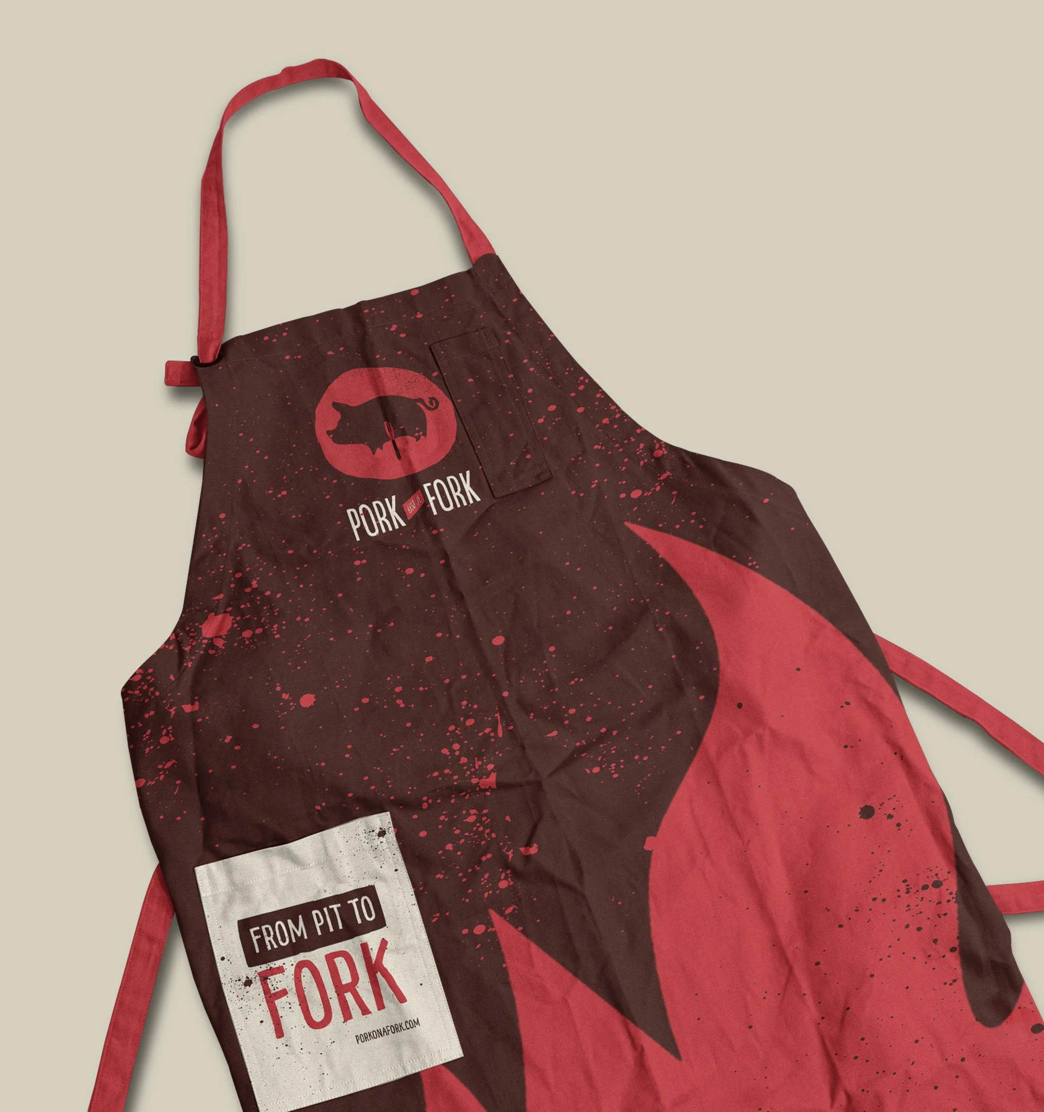 A black apron with red splatters and the Pork on a Fork logo, featuring a pig silhouette on a red circle, hanging against a light beige background.